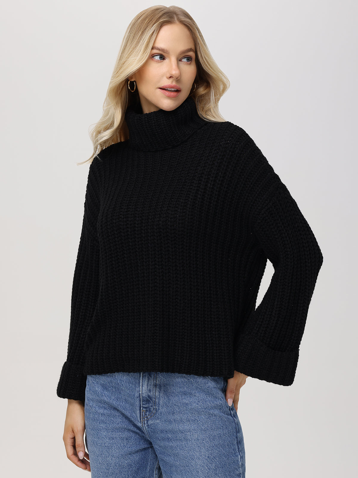 A person with long blonde hair wears a cozy 525 America turtleneck and blue jeans. The oversized sleeves add elegance as they stand against a plain light gray background.