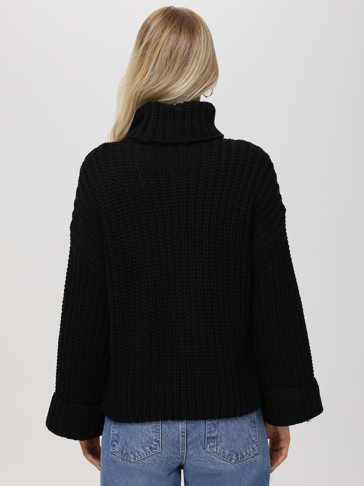 A person with long blond hair is seen from the back wearing a 525 America cozy black turtleneck Shaker Pullover, featuring a chunky textured knit design, oversized sleeves, and rolled cuffs, paired with blue jeans against a plain white background.