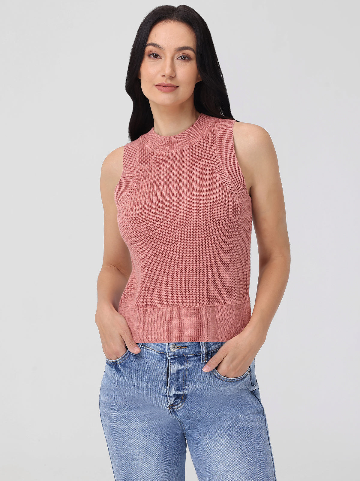 A woman with long black hair showcases chic simplicity in a sleeveless, ribbed, pink knit top by 525 America and light blue jeans, standing with hands in her pockets against a plain white background.