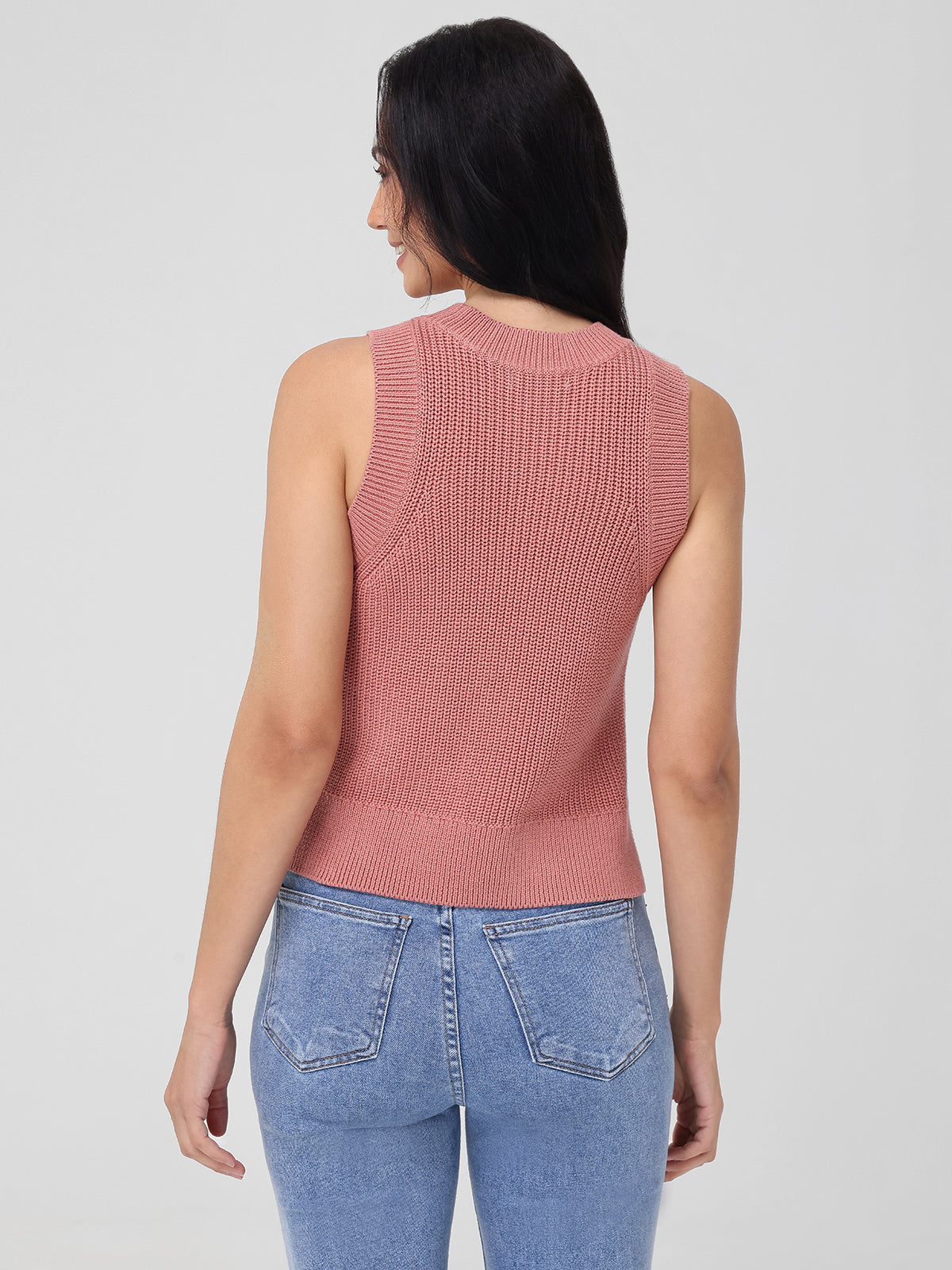 A person with long dark hair stands sideways in a sleeveless pink knit top and light blue jeans from 525 America. Set against a plain light gray background, the 100% cotton outfit exudes chic simplicity as they face right, gazing slightly downward.