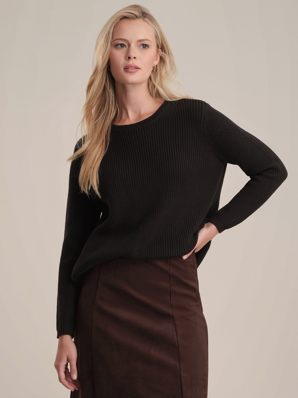 Someone with long blonde hair is wearing a relaxed-fit black knit shaker stitch sweater from 525 America and a dark brown skirt. They strike an effortlessly stylish pose against a light-colored background, hand on hip.