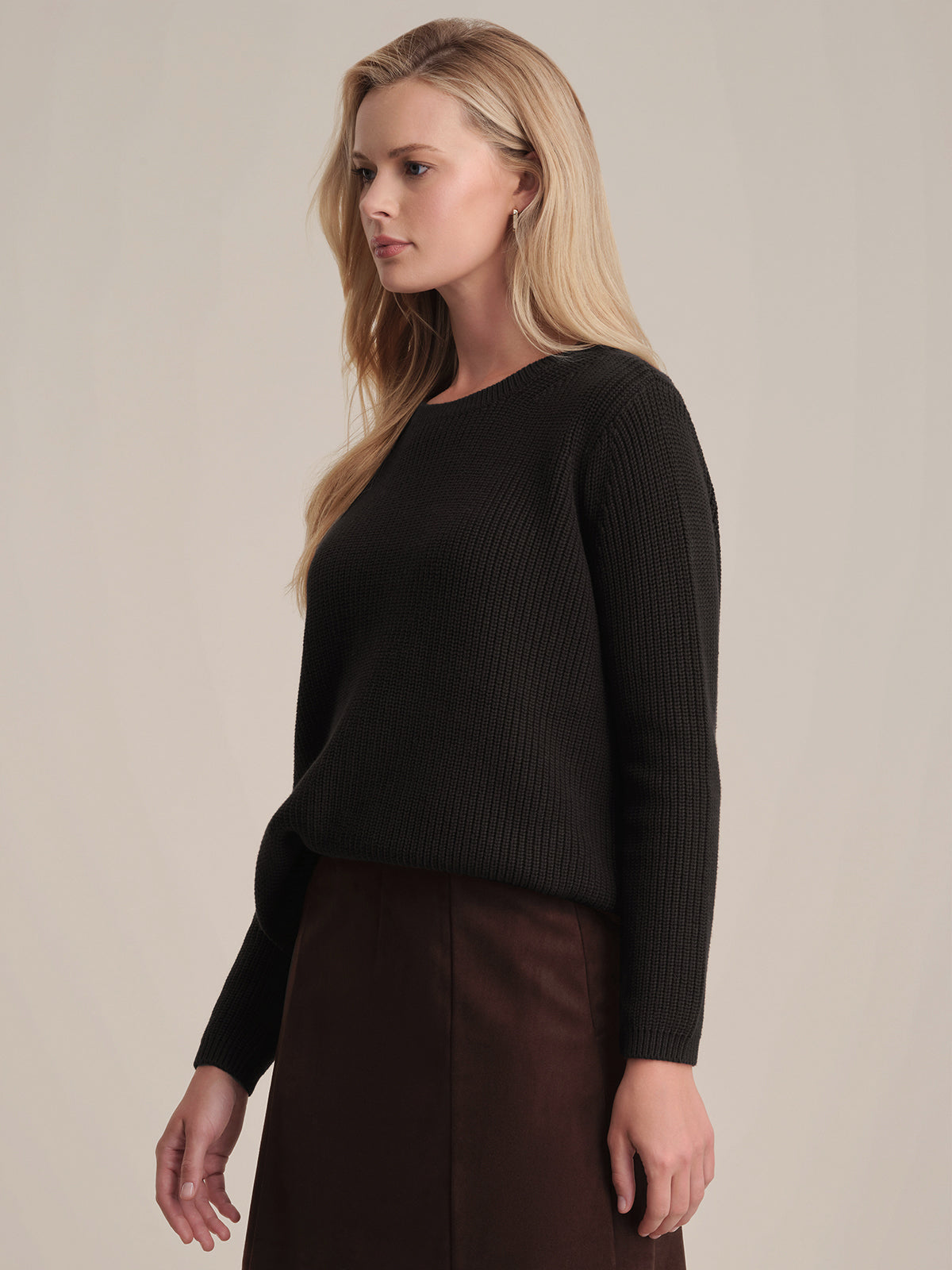 A person with long blonde hair poses in a black shaker stitch sweater by 525 America, made from 100% cotton, featuring a relaxed fit. They pair it with a brown skirt and stand against a neutral backdrop, gazing left.