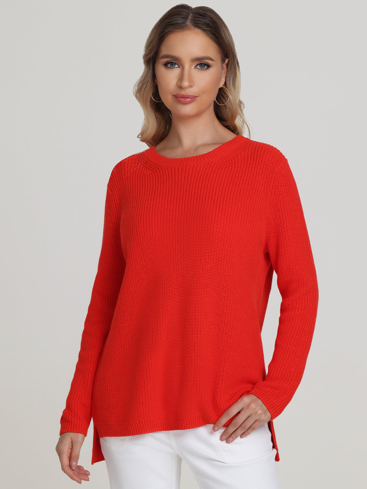 Against a plain background, a woman with shoulder-length hair models versatile style in a bright red shaker stitch sweater and white pants from 525 America, left hand on her hip and gazing directly at the camera.