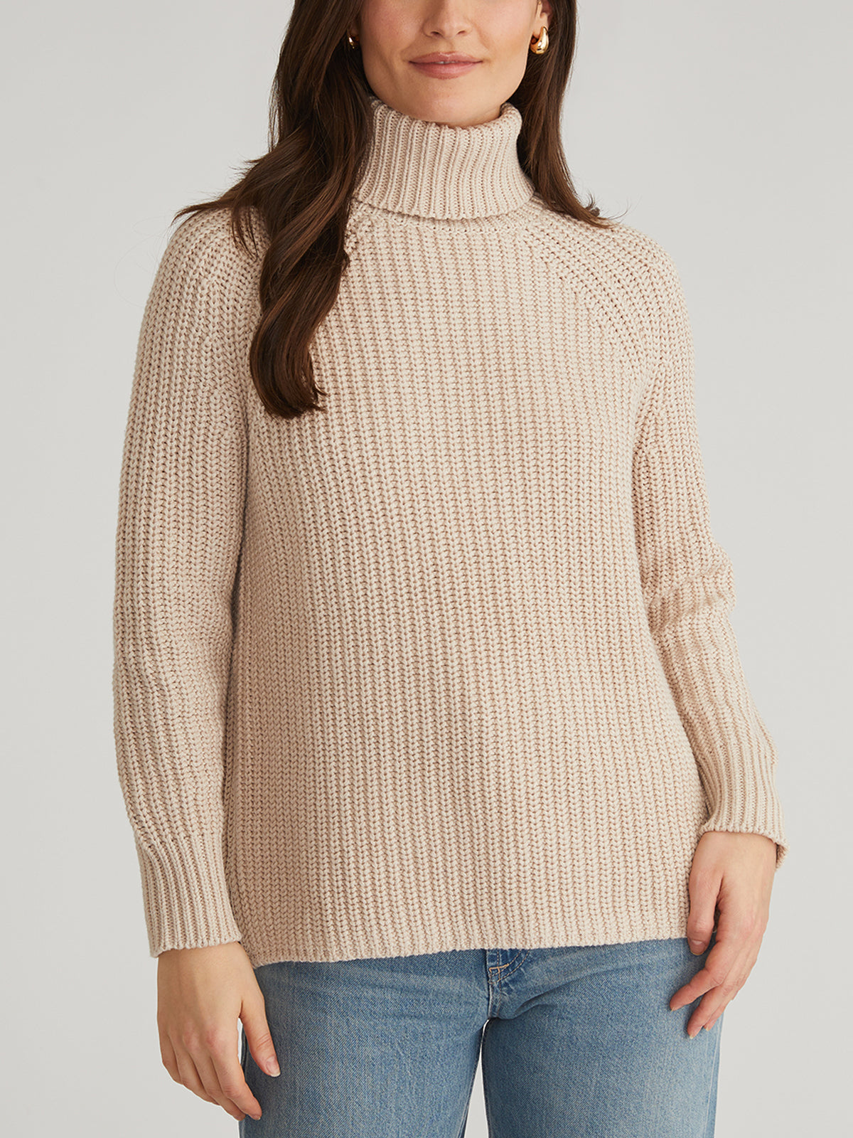 A woman with long brown hair stands against a plain background wearing a cozy 525 America cream-colored turtleneck sweater made of 100% cotton, featuring ribbed cuffs and hem, paired with blue jeans. Her hands rest casually by her sides.