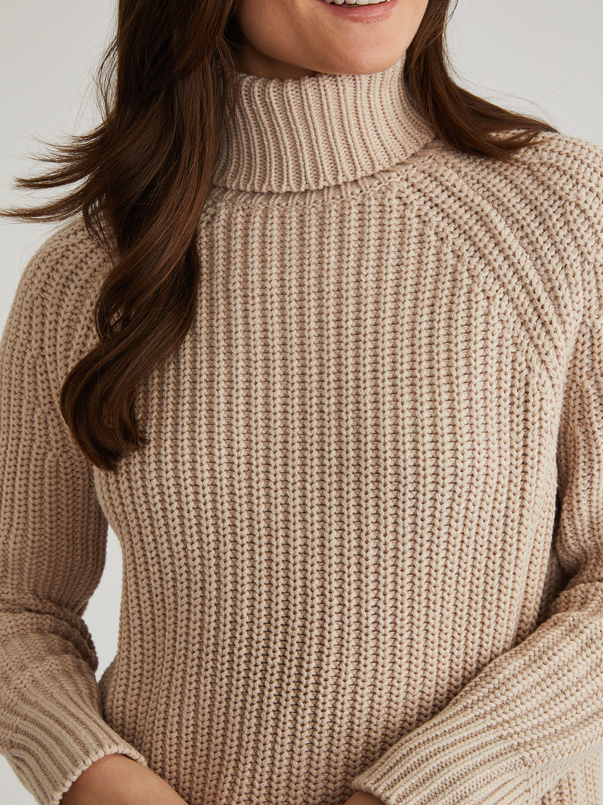 Someone is wearing a cozy, beige, 525 America turtleneck sweater made of 100% cotton with a textured design and long sleeves featuring a ribbed pattern. Their long, brown hair flows over their shoulders against a plain, neutral background.