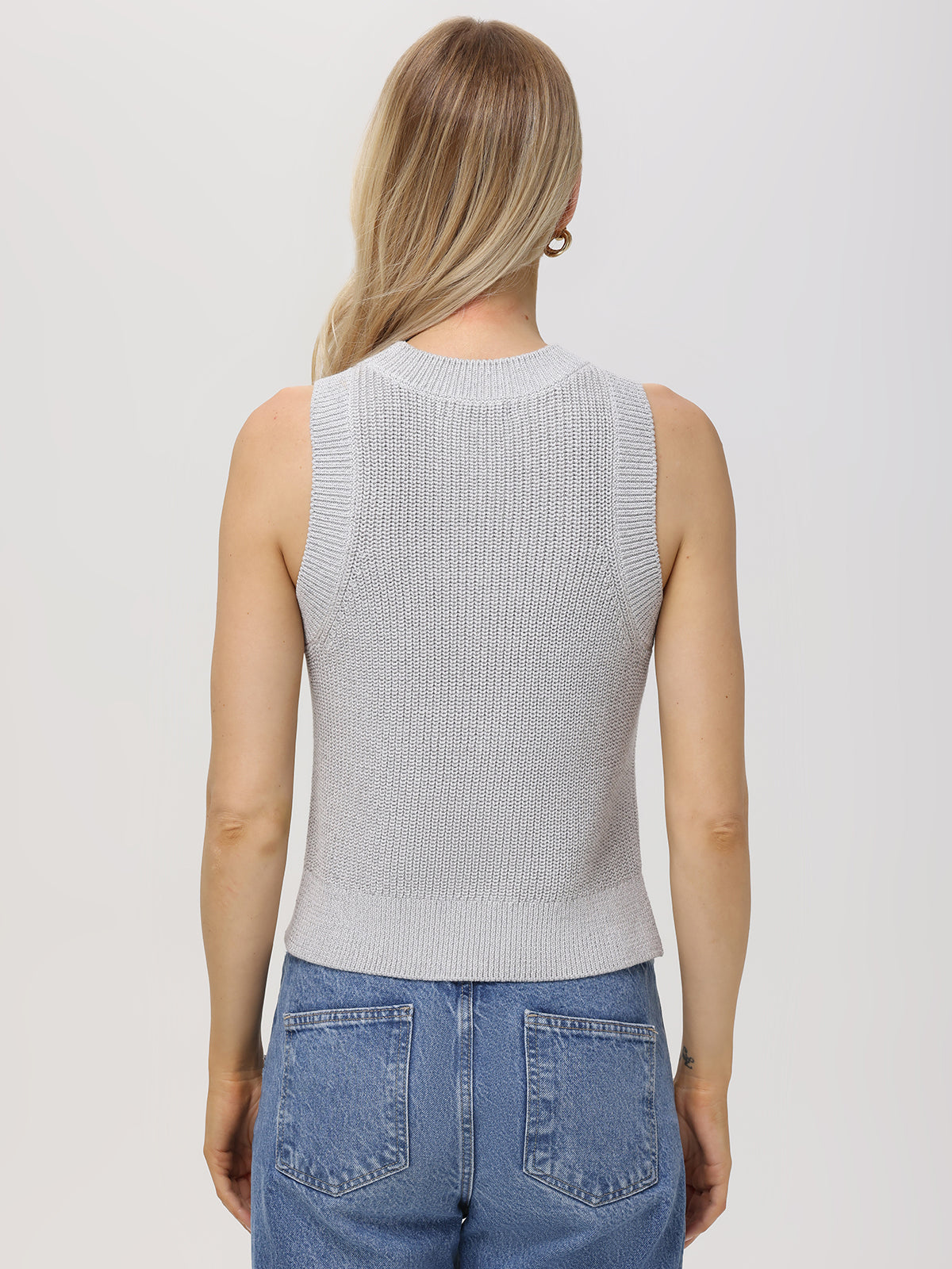 A person with long blonde hair faces away in a dazzling outfit, featuring a 525 America light gray fitted sleeveless knit top and blue jeans. The plain white background accentuates the ensembles subtle elegance.