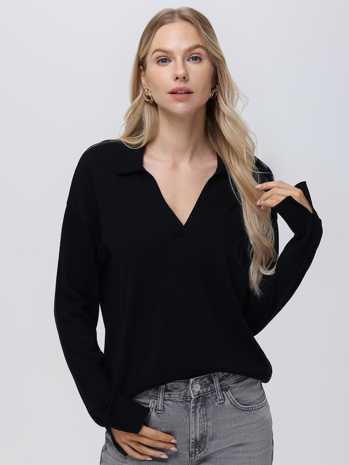 A woman with long blonde hair wears a black lightweight 525 America pullover featuring a stylish Johnny collar, paired with light gray jeans. She stands against a plain white background, looking directly at the camera with one hand slightly raised.