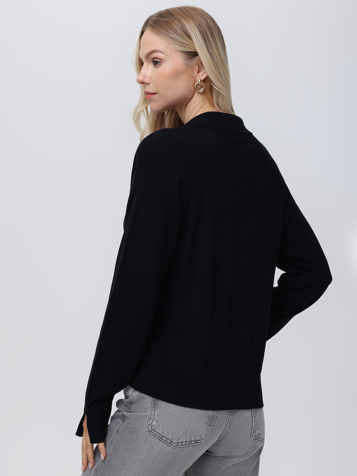 A person with long blonde hair stands sideways, wearing a black Johnny collar sweater by 525 America, paired with gray jeans. The lightweight pullover offers breathable comfort against the plain, light-colored background.