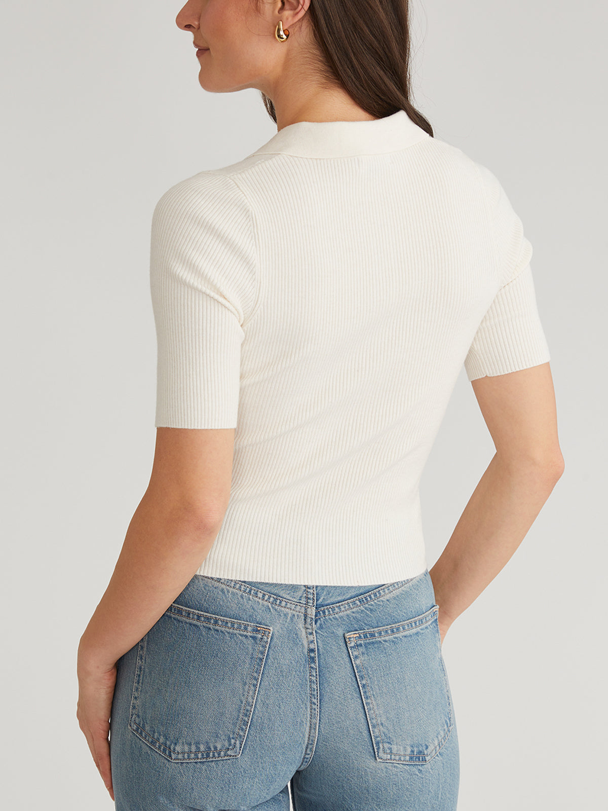 A person stands with their back to the camera, wearing a 525 America top featuring a rib design and Johnny collar. Paired with blue jeans, they have medium-length brown hair and gold hoop earrings against a neutral background.