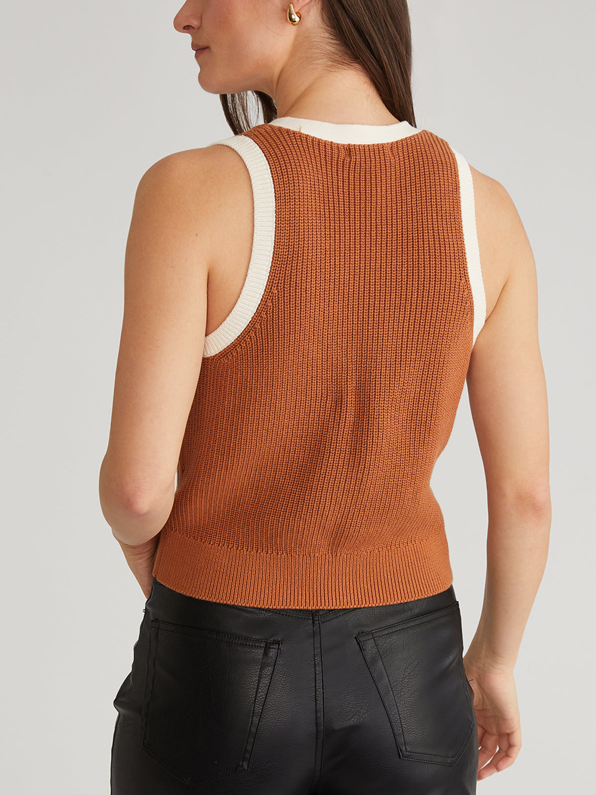 Rear view of a person in a Claudia Tipped Button Up Vest, an orange sleeveless knit with cream trim, paired with black leather pants. The long-haired individual is shown waist-up, creating a sophisticated layered look.