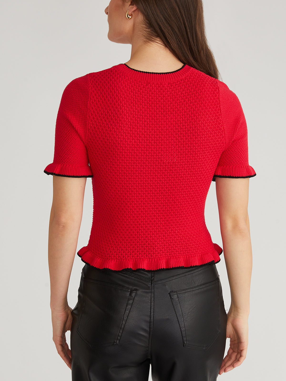 A person with long brown hair exudes sophistication in a 525 America mixed-stitch red short-sleeve top, featuring black trim and ruffles, seamlessly paired with high-waisted black leather pants against a plain, light background that highlights the chic ensemble.