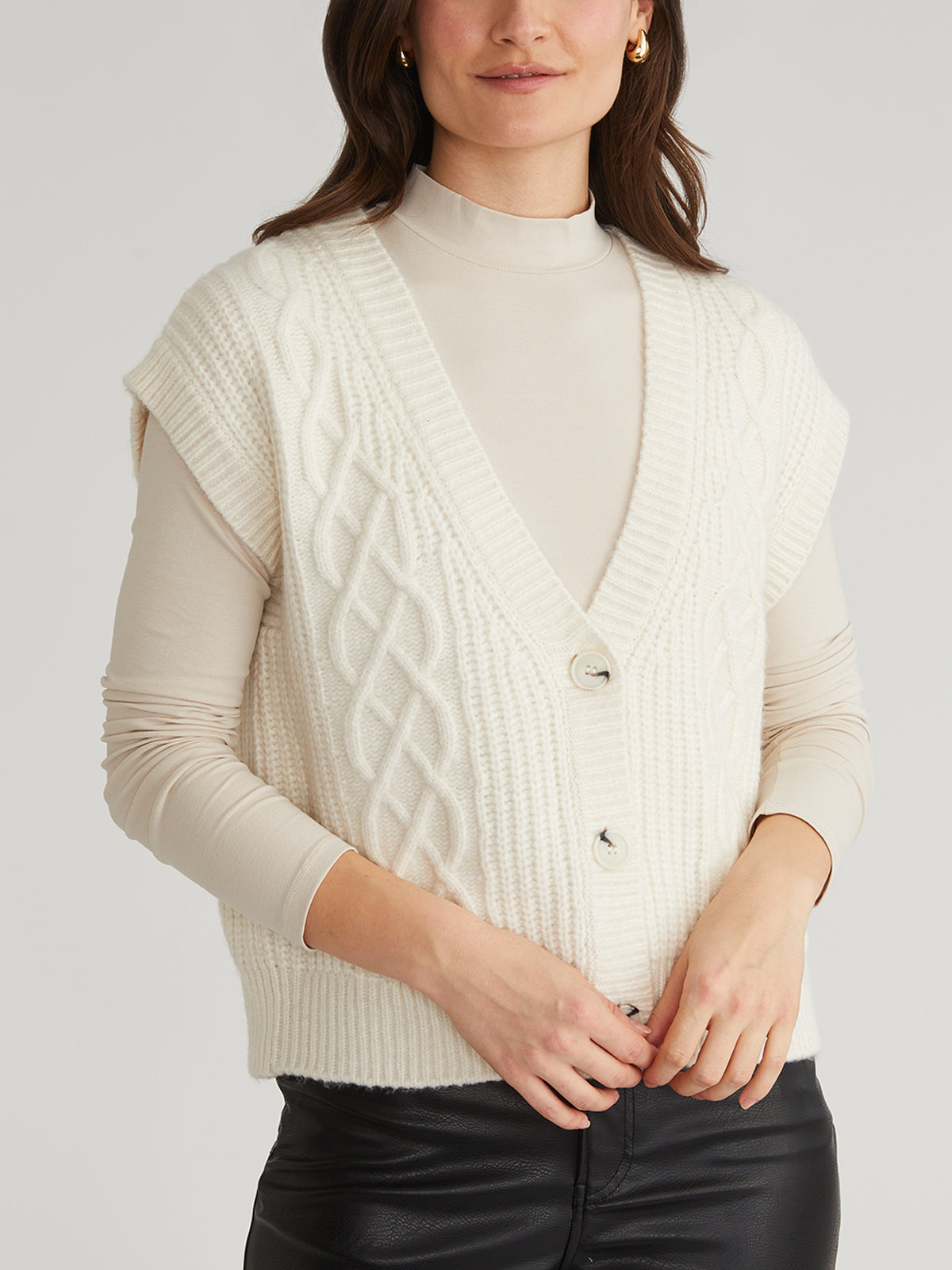 Dressed in a chic ensemble, a person wears a cream V-neck button front vest by 525 America over a beige long-sleeve turtleneck top and black pants, offering an elegant twist to the classic cable sweater look.