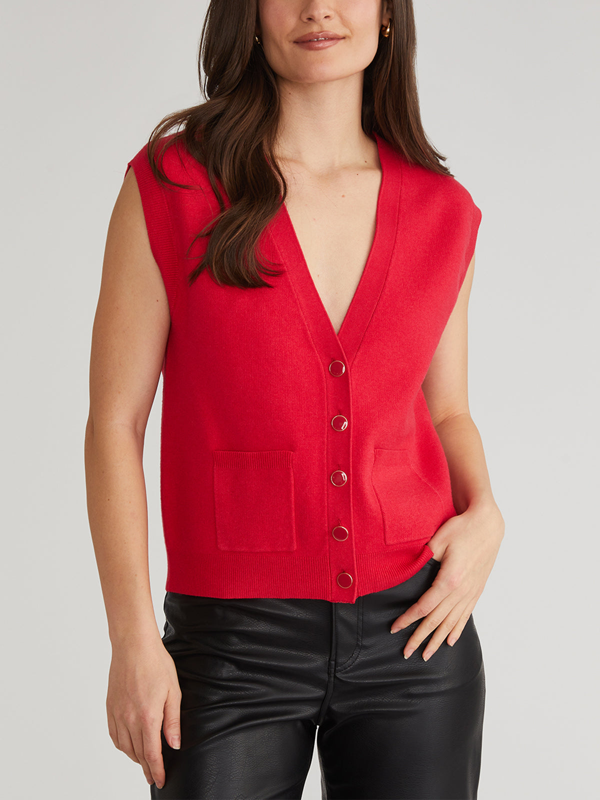 A person elegantly poses against a light gray background, showcasing perfect layering in a stylish button-down sleeveless red vest with pockets by 525 America, paired seamlessly with black leather pants. The individual has long brown hair.