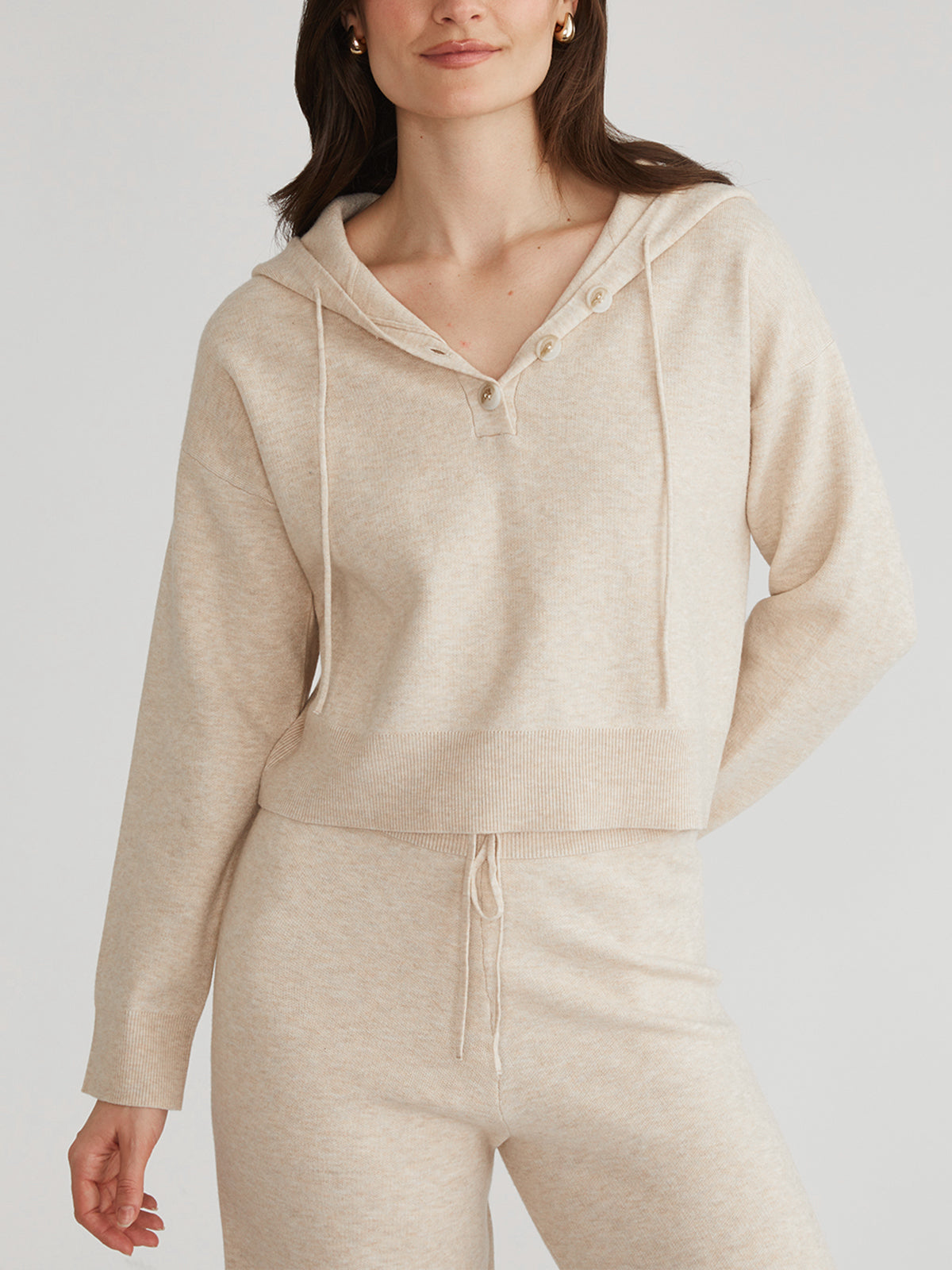 A person in a cozy, relaxed fit beige V-neck sweater with hood by 525 America, featuring drawstrings and matching pants, stands against a neutral background with hands by their sides and long hair flowing.