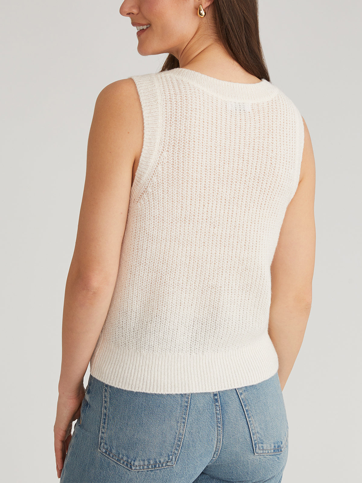 A woman with long brown hair, in a sleeveless white knit top by 525 America featuring pointelle stitching and ribbed texture, paired with blue jeans and gold hoop earrings, is shown from the back against a plain, neutral background that enhances her elegant look.