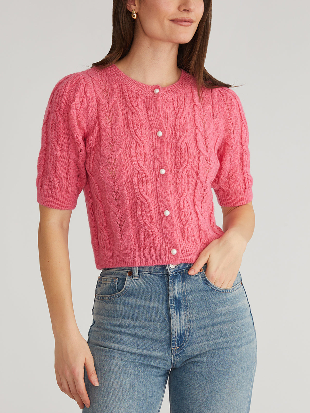 A person wears a 525 America pink cable-knit crewneck cardigan with puff short sleeves and pearl-like buttons, paired with blue high-waisted jeans against a plain, light-colored background.