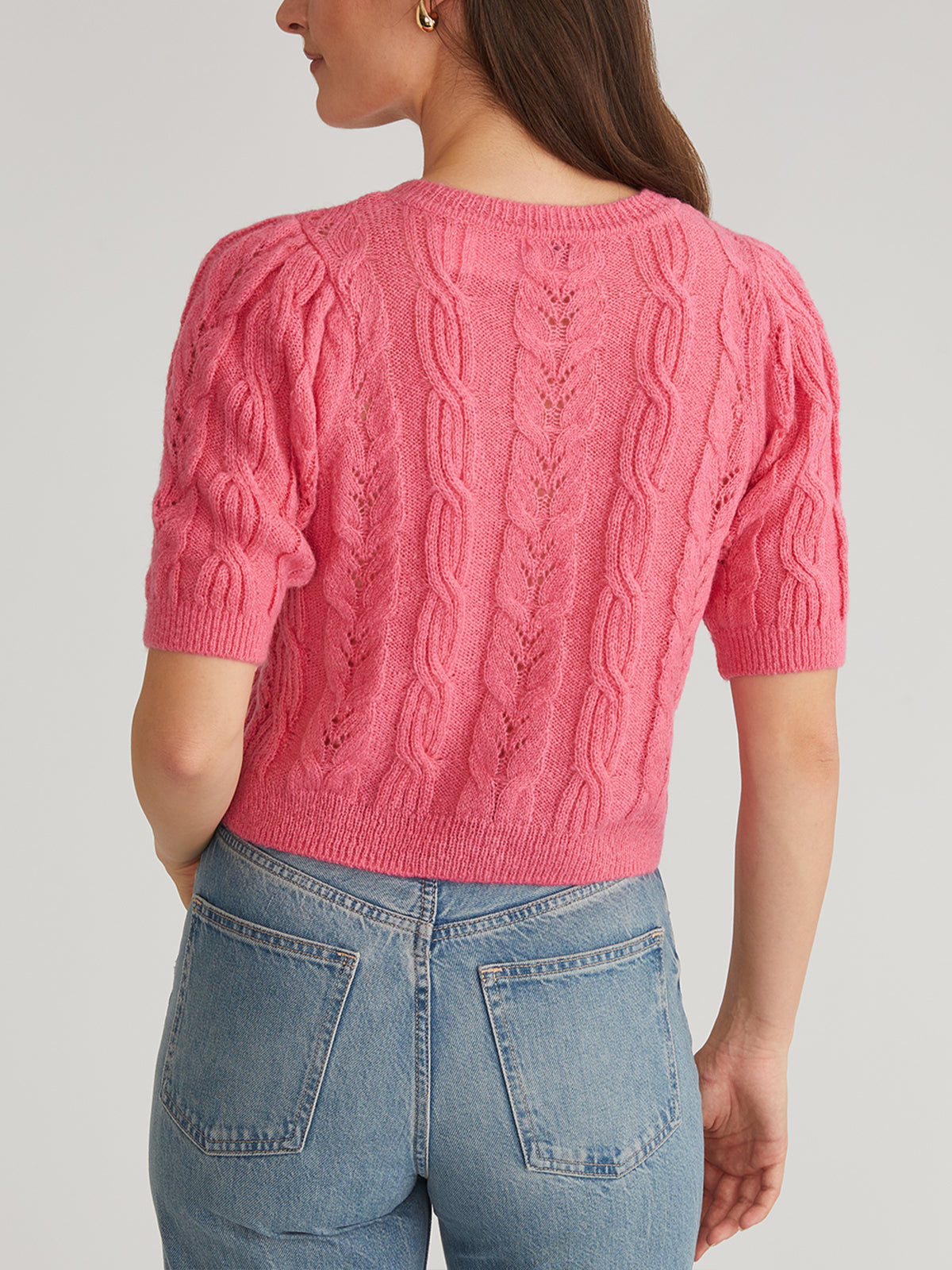 Wearing a pink cable-knit short-sleeve sweater with puff sleeves by 525 America, someone is facing away from the camera. They pair it with light blue jeans against a plain gray background.