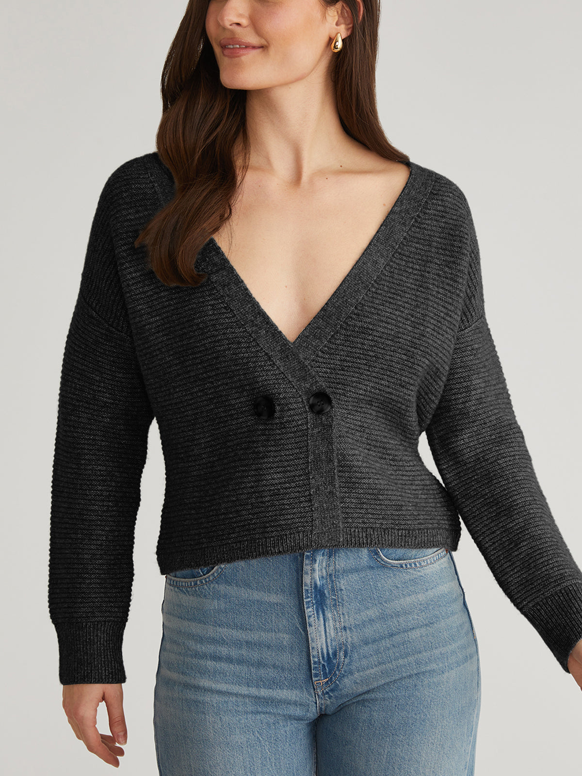 Wearing a dark gray, textured ottoman stitch cardigan from 525 America with a deep V-neck and two buttons, paired with light blue jeans, a woman stands against a plain background. Her hair is partially visible as it falls over her shoulder.