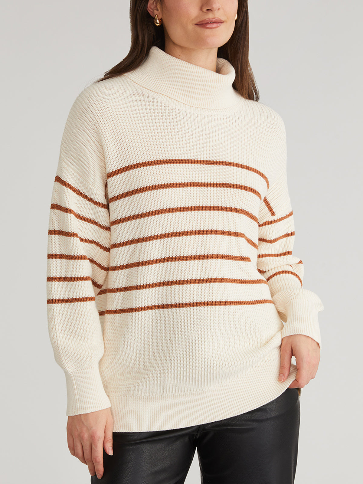 A person is stylishly dressed in a 525 America oversized striped turtleneck pullover sweater, made of 100% cotton featuring a cream color with brown stripes, paired with black pants. Their hair is down, and one hand is casually in their pocket against a plain gray backdrop.