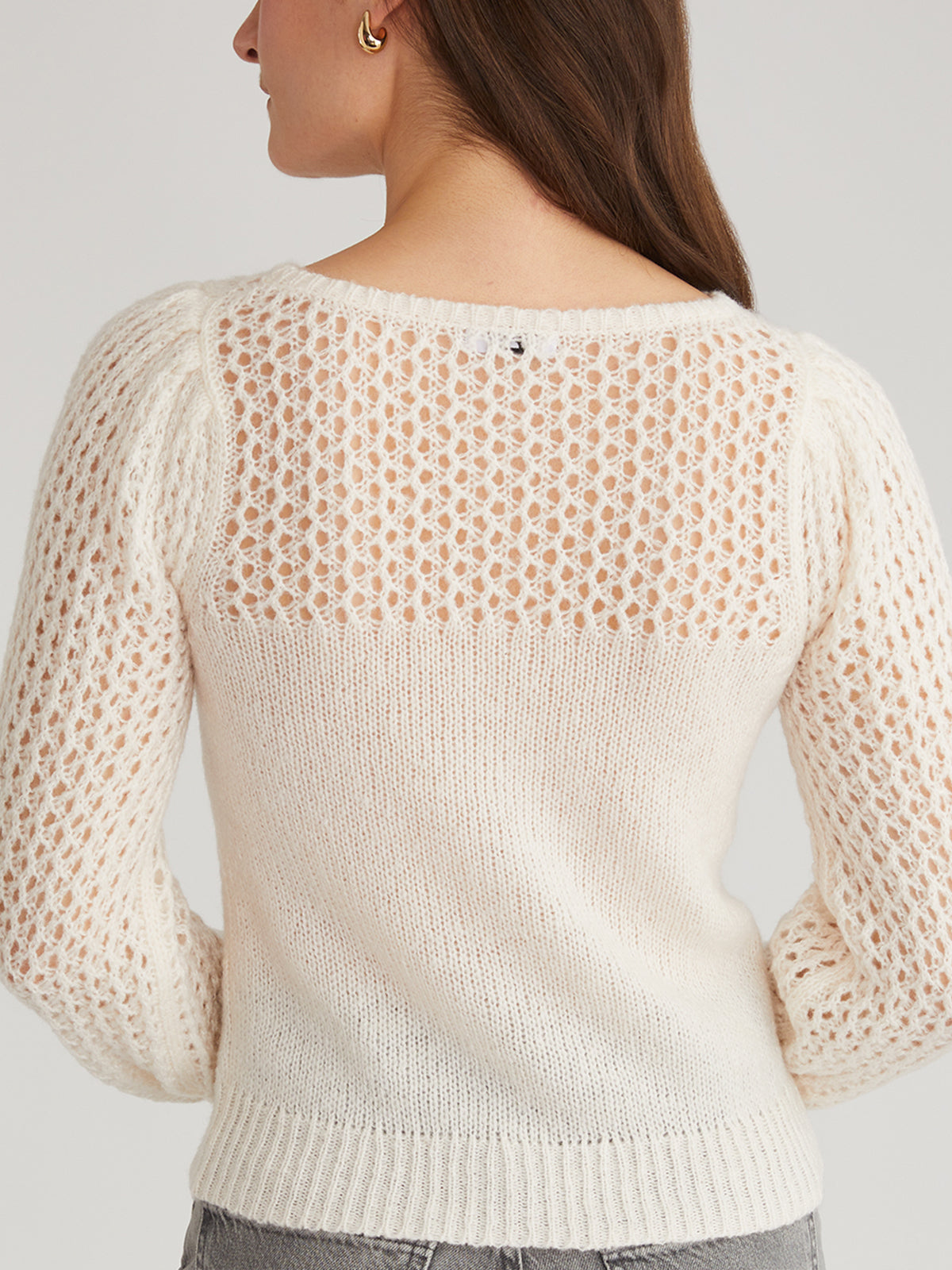 A person models a cream 525 America long-sleeve sweater with lace-knit back and puff sleeves, paired with gray pants, set against a plain light backdrop. Their shoulder-length brown hair enhances the modern look.