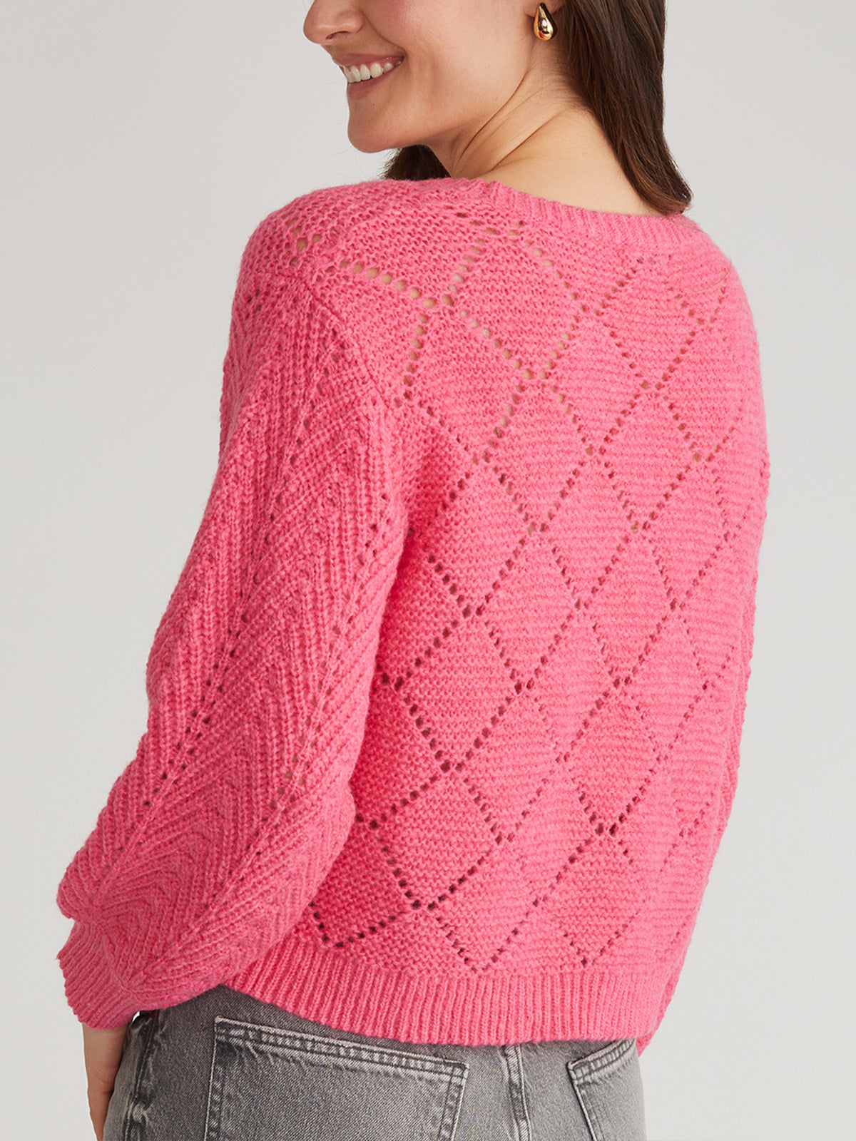 A person in a vibrant pink pointelle pullover by 525 America, featuring a diamond pattern and textured balloon sleeves, stands with their back slightly turned, smiling. They wear gray jeans and have long hair down.