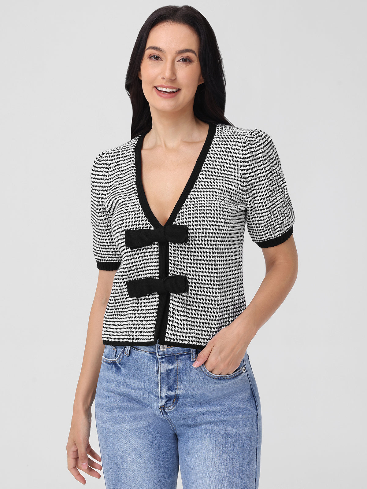 A person with long dark hair smiles at the camera, exuding feminine charm in a V-neck Elaine Puff Sleeve Tie Front Top with black and white patterns and black trim, paired with blue jeans—a perfect wardrobe addition known for its effortless style.