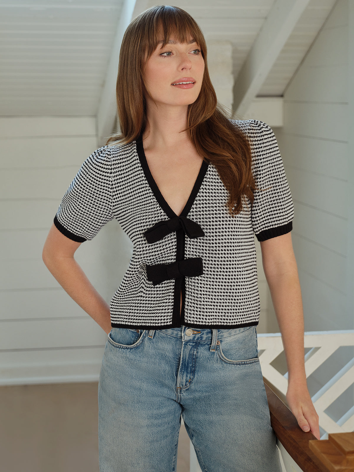 Indoors, a person with long hair leans on a wooden railing, showcasing the Elaine Puff Sleeve Tie Front Top with black accents and light blue jeans, elegantly layered with a chic cardigan. The bright and airy space features white walls and a sloped ceiling, enhancing the feminine aesthetic.