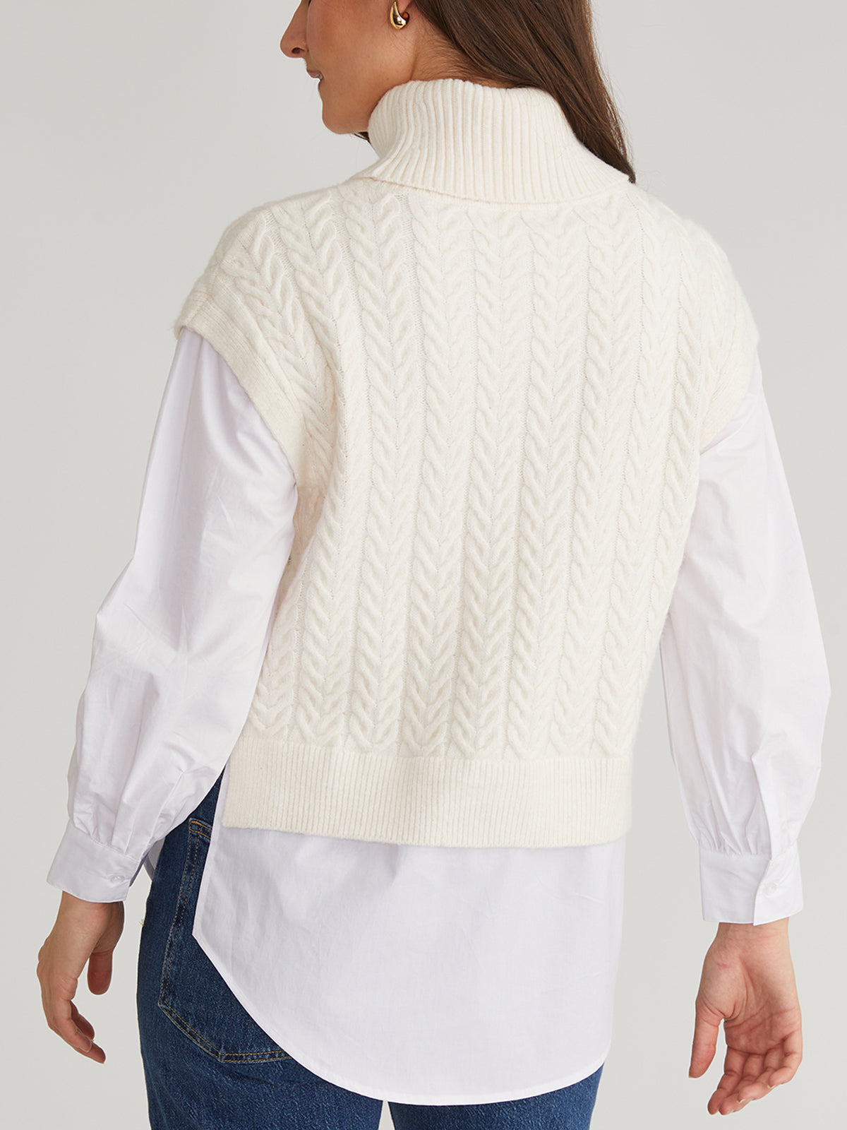 A person is wearing a cozy, white cable-knit turtleneck sweater by 525 America over a white long-sleeve shirt with blue jeans, viewed from the back with their head slightly turned to the right.