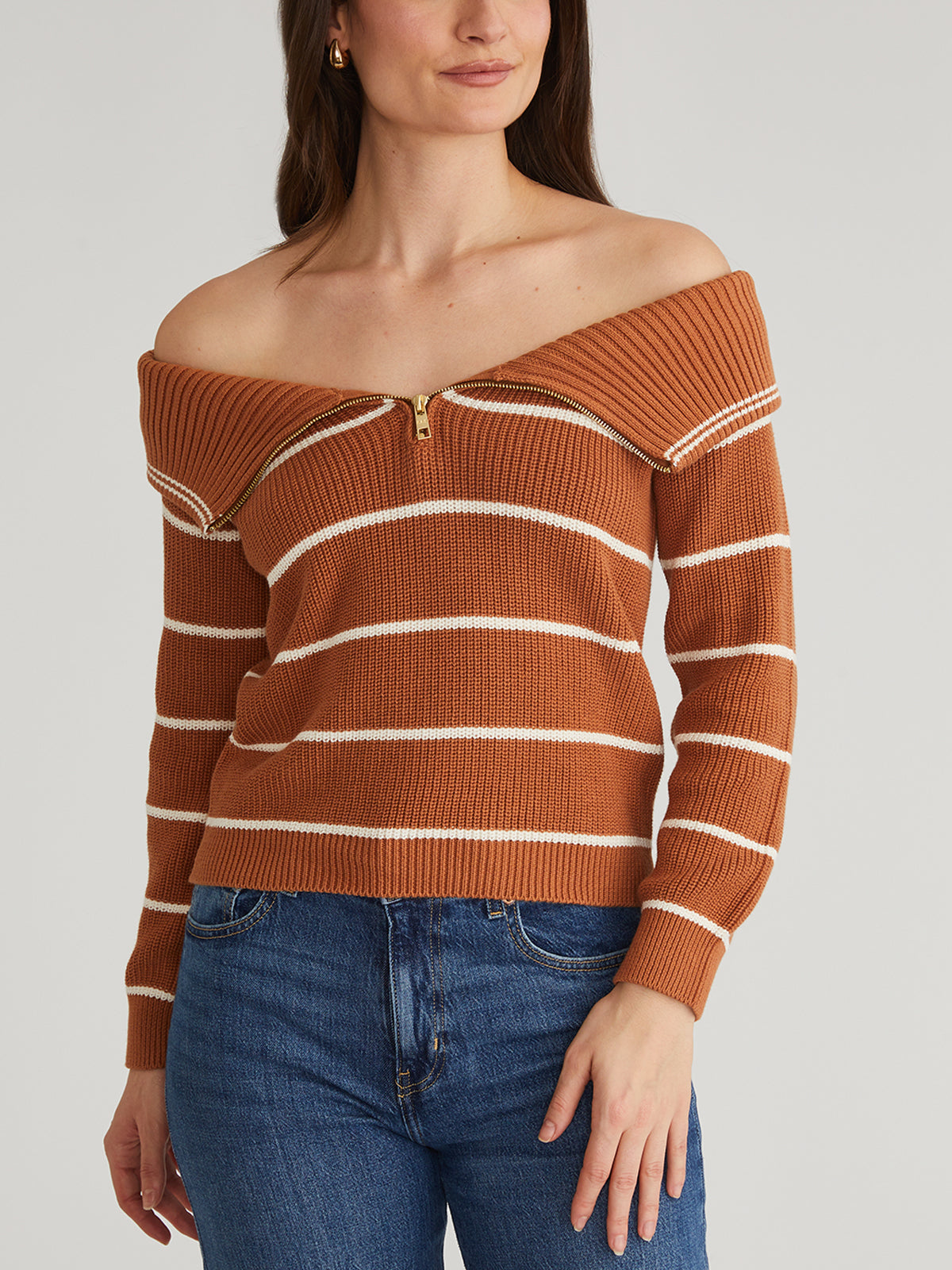 A person is modeling a brown, 100% cotton off-the-shoulder sweater with white stripes and an adjustable zippered neckline by 525 America, paired with blue jeans. The model has long hair, posing with one hand on their hip against a plain gray background.