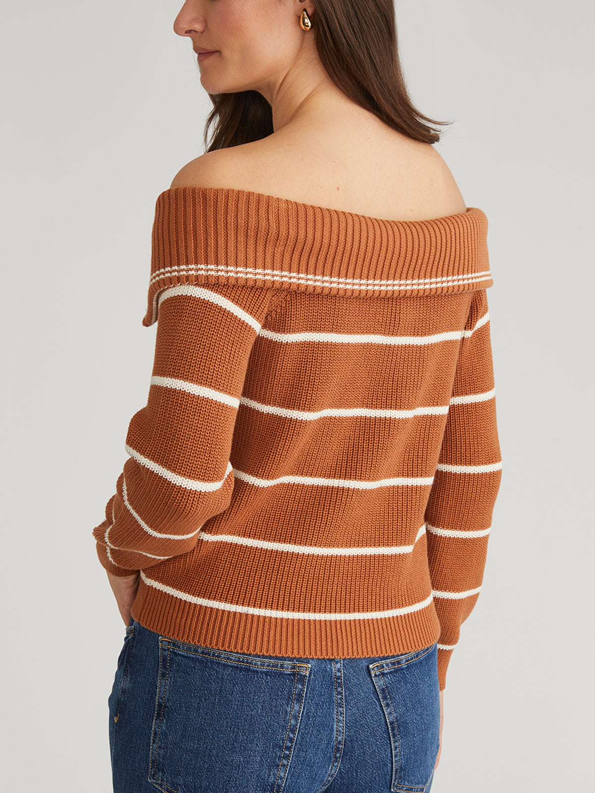 The person is wearing a 525 America off-the-shoulder, brown and white striped pullover made of 100% cotton. With their back to the camera and hands tucked into blue jeans, they exude effortless style.