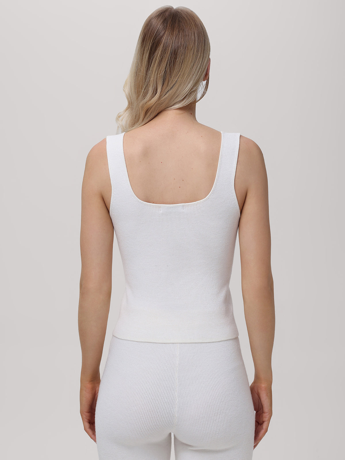 A person with long, light hair is shown from behind wearing a versatile white sleeveless top and matching pants from 525 America. The form-fitting outfit stands out against the plain gray background.