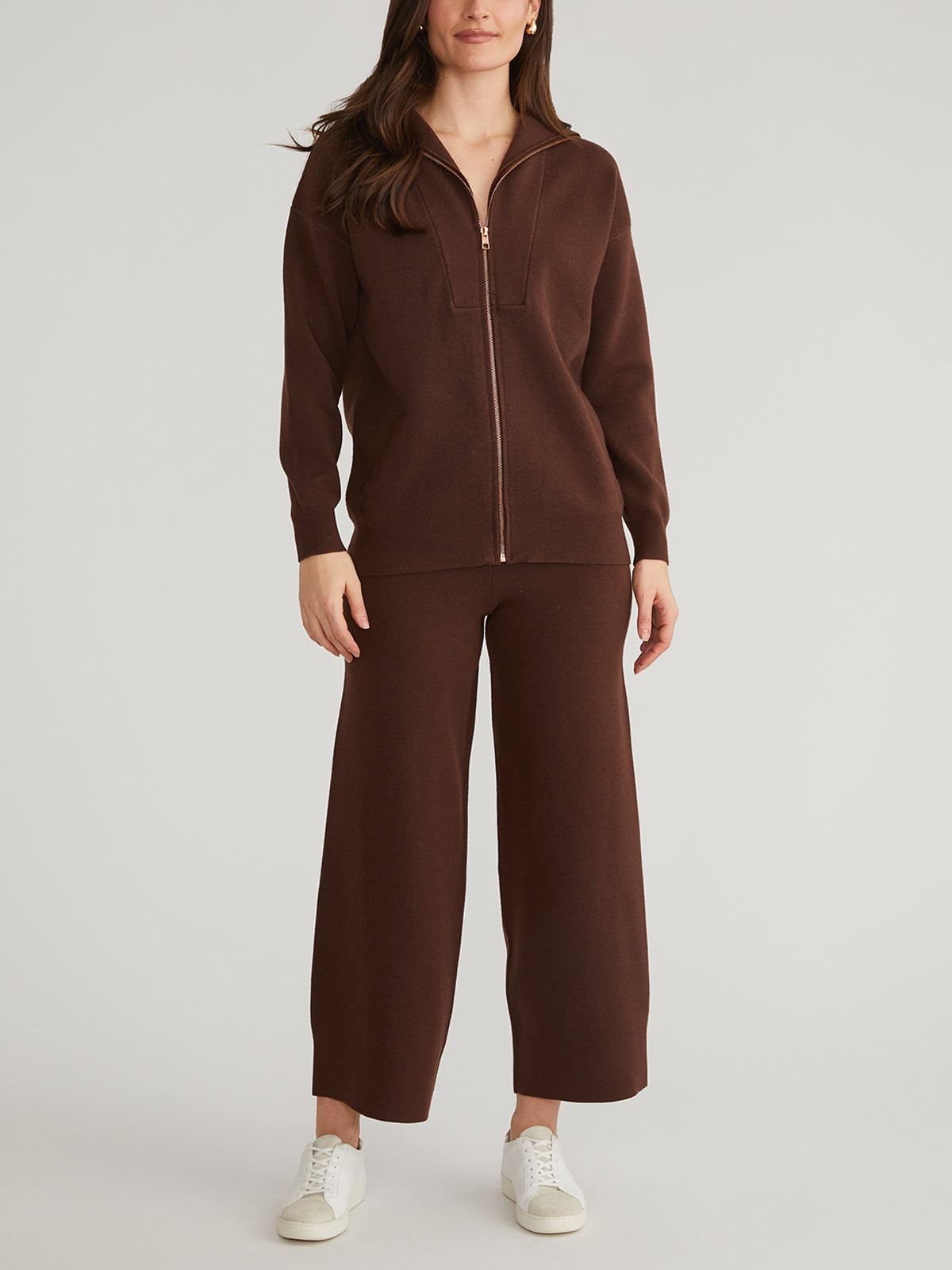 A person wears a matching brown zip-up jacket and high-rise wide-leg pants from 525 America, with their long hair cascading down. They pair the outfit with white sneakers against a plain, light backdrop that highlights the elegance of the wide-leg silhouette.