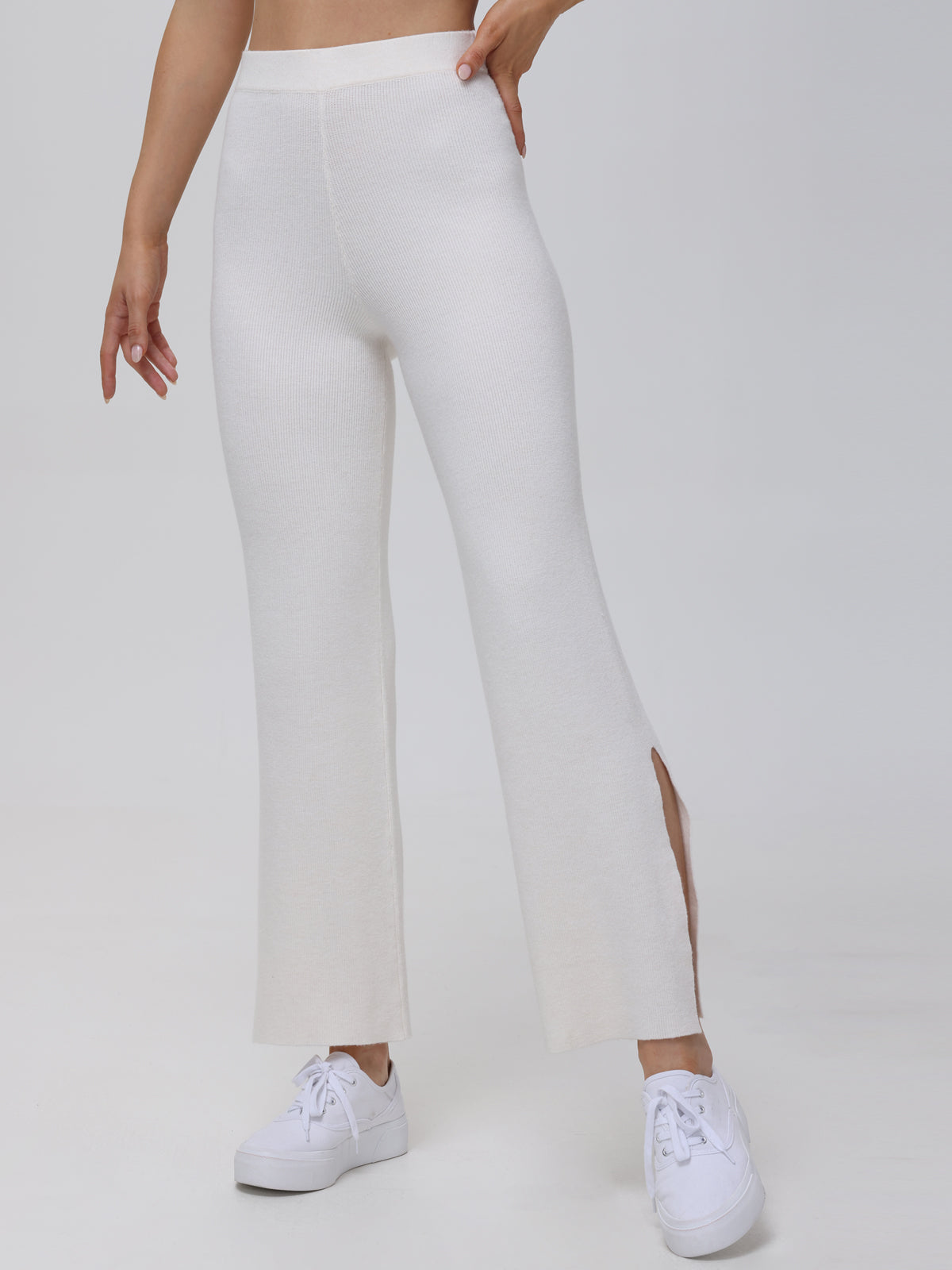 A person sports 525 Americas cream-colored knitted trousers with a flared leg and subtle side slits, effortlessly paired with white sneakers. Their hand rests on the hip against a plain light gray backdrop, enhancing the outfits understated elegance.