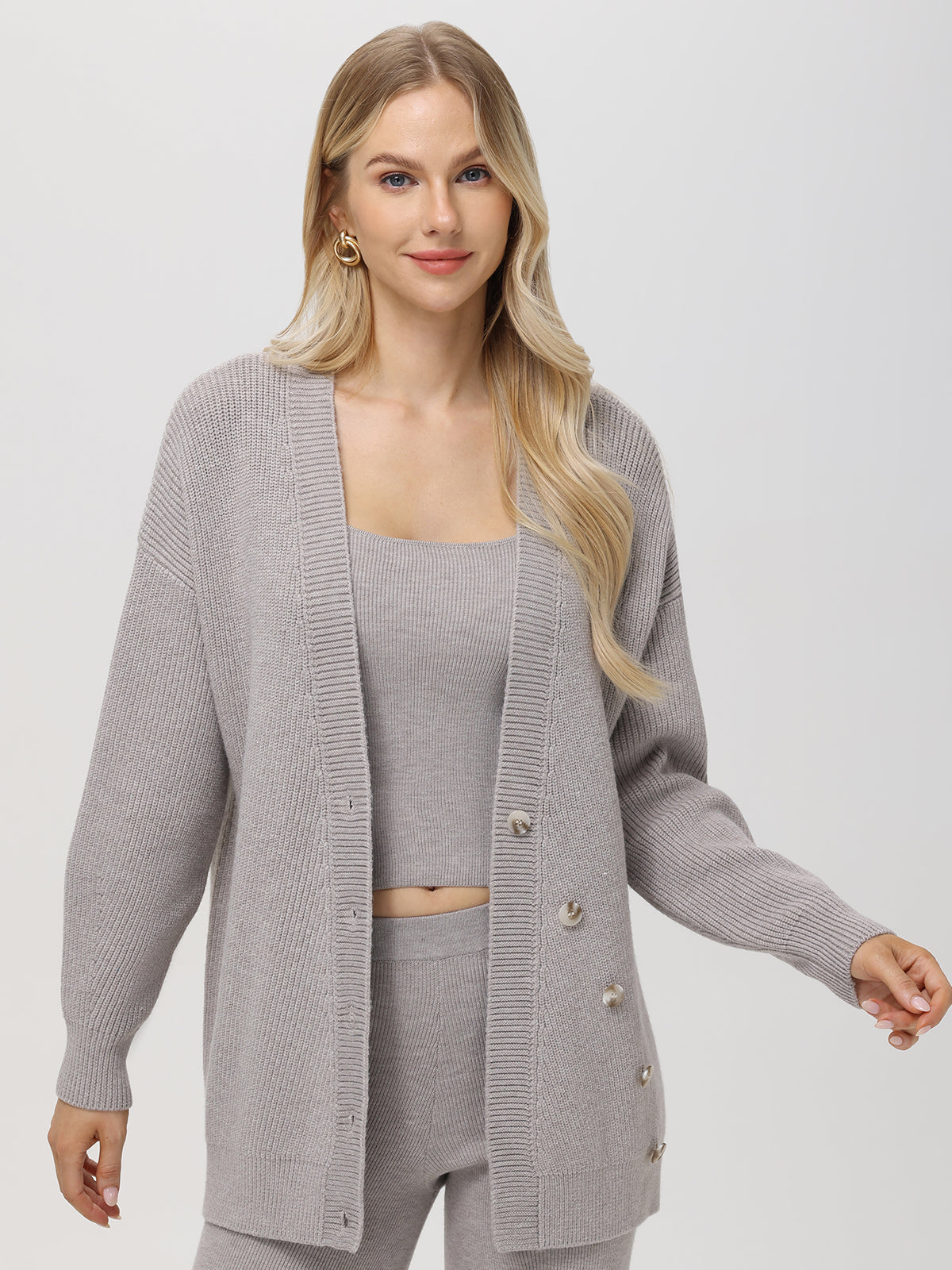 A person with long hair sports a light gray, ribbed knit cardigan with pearl buttons over a matching top from 525 America. The relaxed fit enhances their style as they stand against a plain, light-colored background.