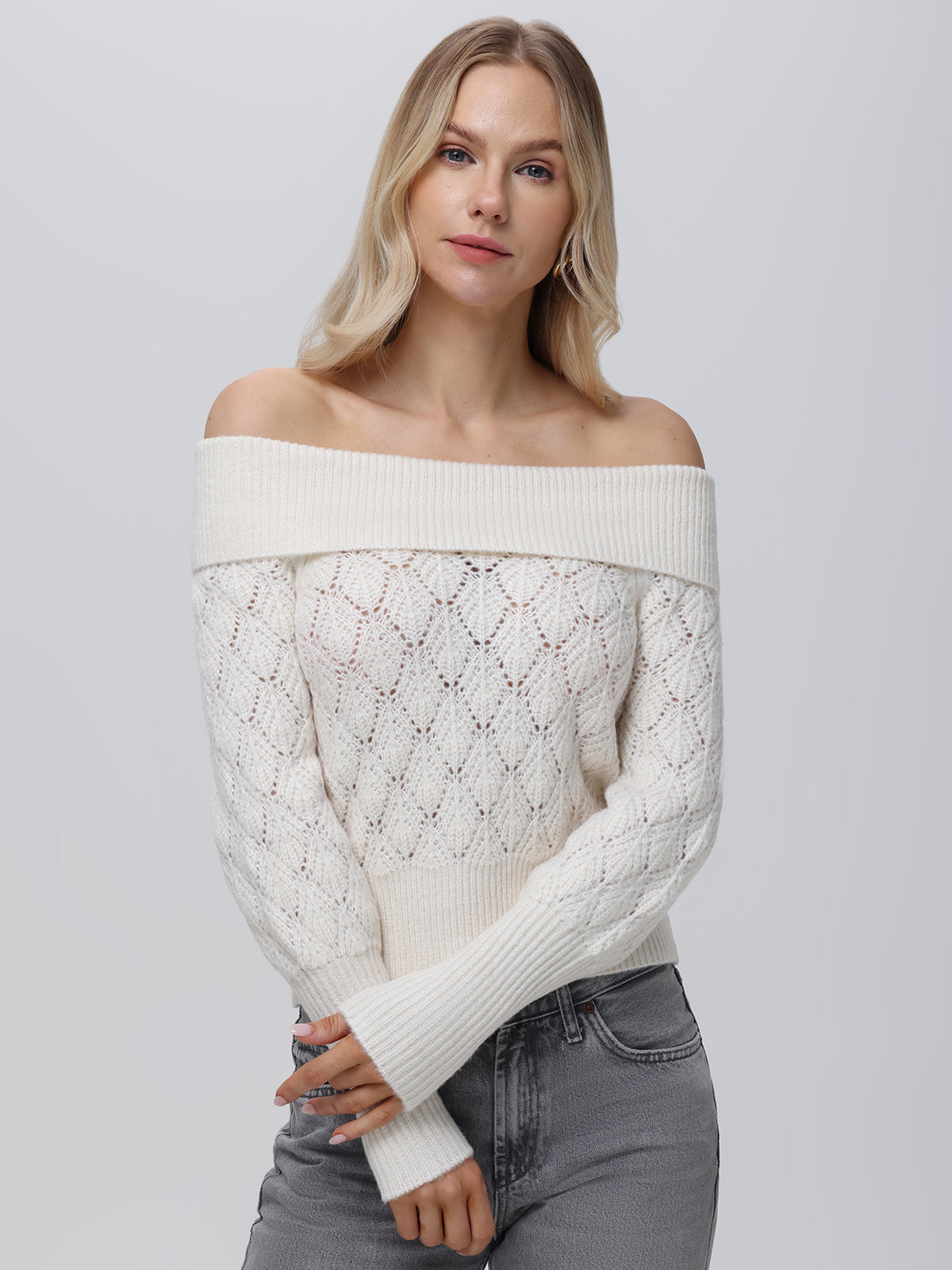 A woman with long blonde hair wears a 525 America off-the-shoulder white knit sweater and gray pants. Posed against a plain background, she looks directly at the camera with gently crossed arms, highlighting this versatile wardrobe essential.