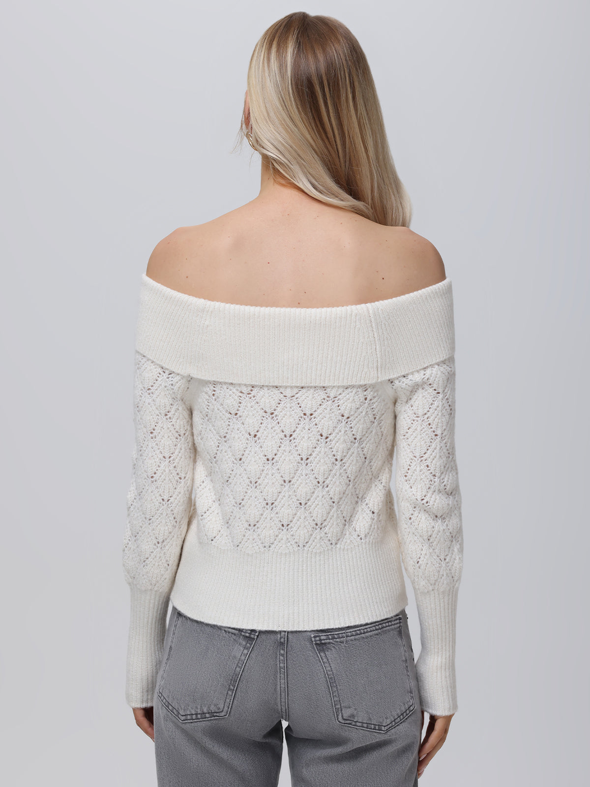 A person with long blonde hair is wearing a 525 America white, off-the-shoulder Pointelle pullover with a textured diamond pattern and faded gray jeans, captured from the back against a plain background.