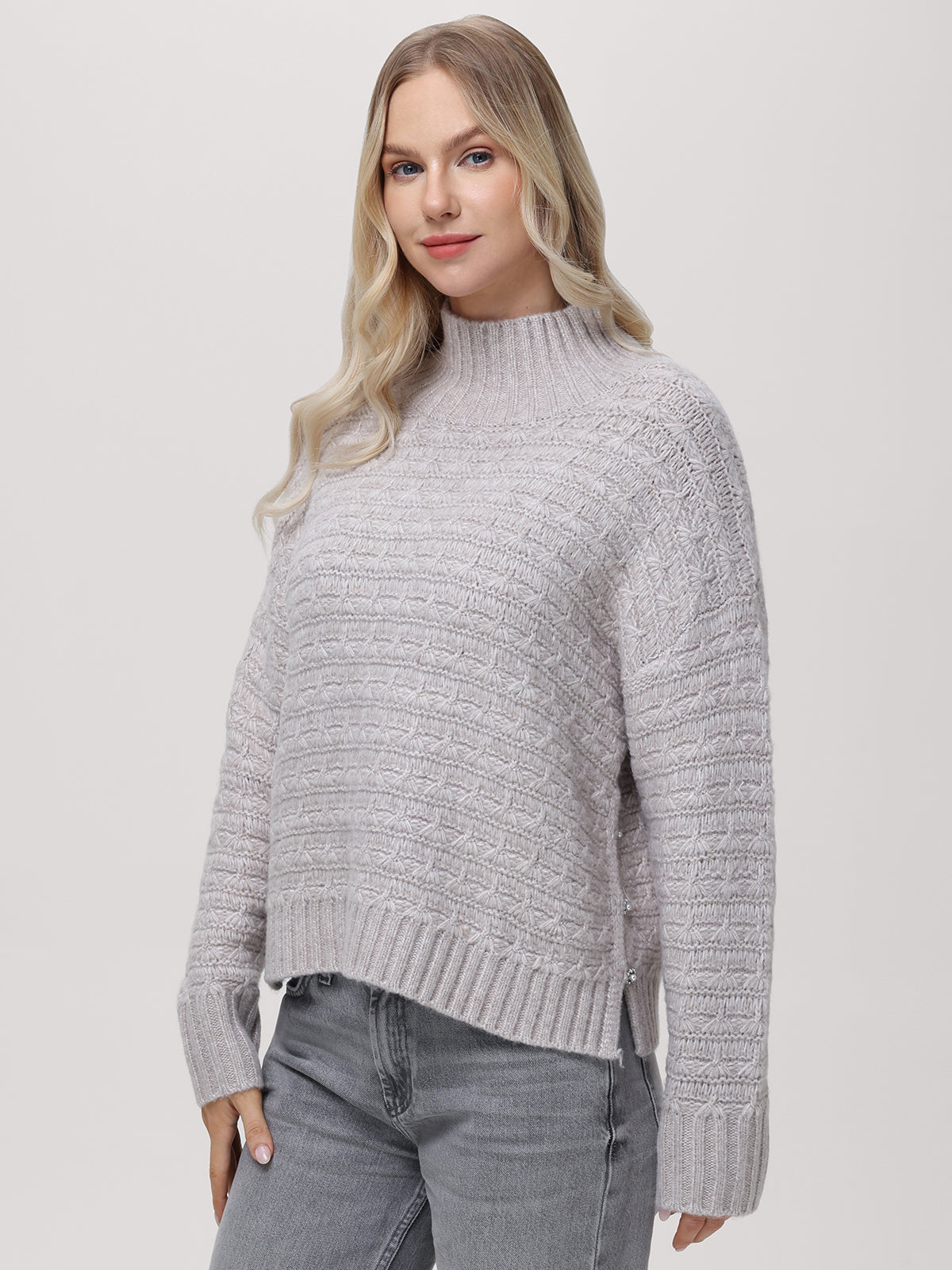 Against a light gray background, a person with long, wavy blonde hair wears a 525 Americas Layla Mockneck Pullover featuring button accents and pairs it with gray jeans. They gaze at the camera with a neutral expression.