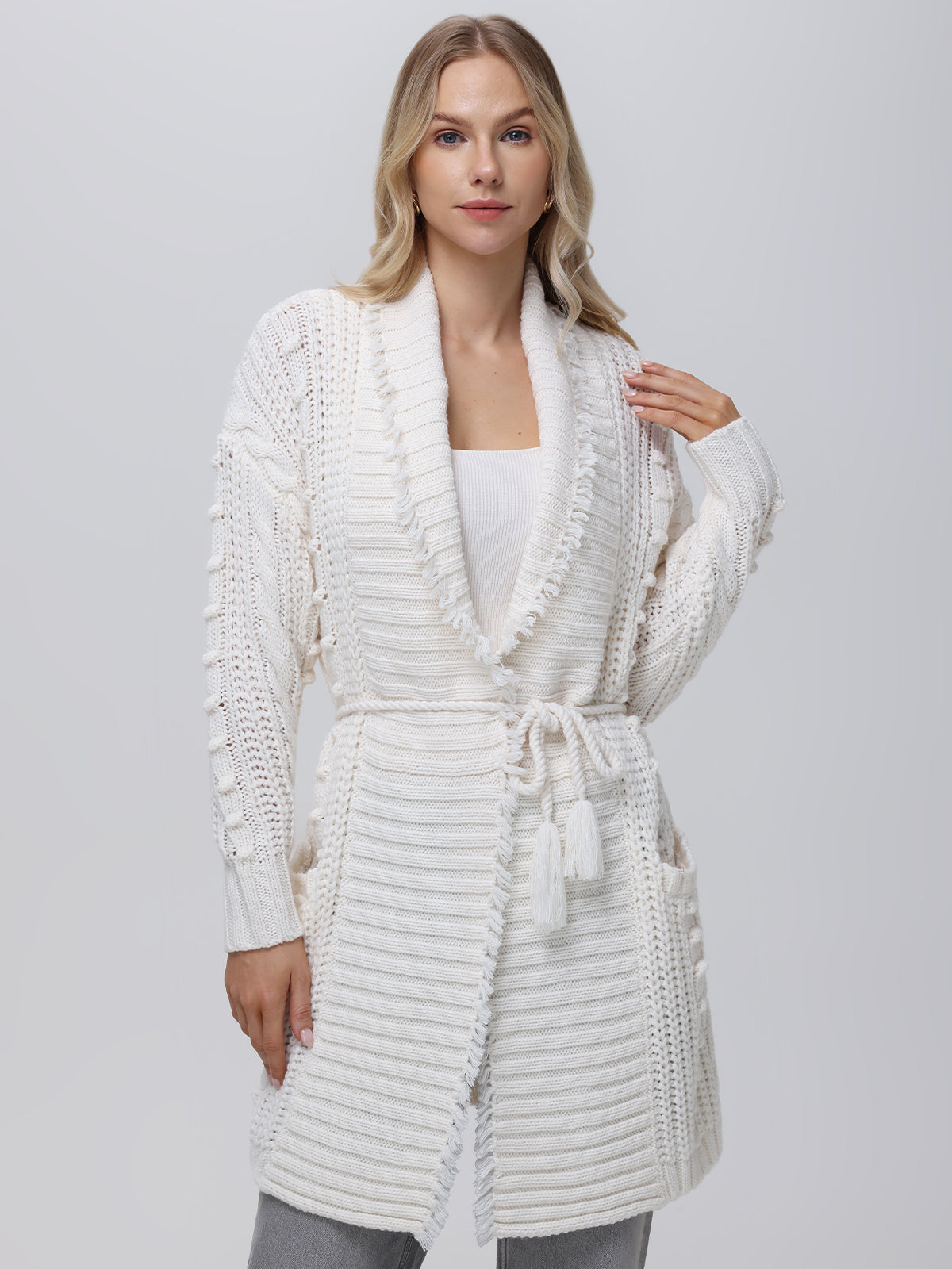 Styled in sophistication, a person wears the Penelope Cable Sweater Coat by 525 America—a white, knitted cardigan with textured patterns, pockets, and matching belt—over a white top and gray pants against a light gray background.