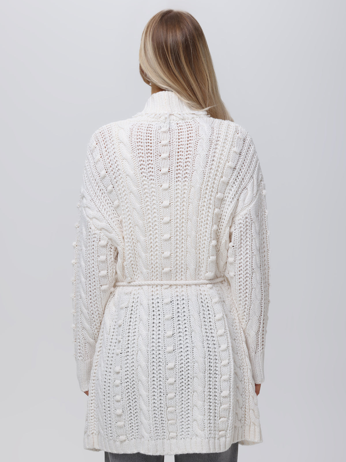 A person with long hair wears a white knit cardigan with a tied belt, seen from the back. The stylish Penelope Cable Sweater Coat by 525 America shows off its cable and bobble patterns against a plain light gray background.