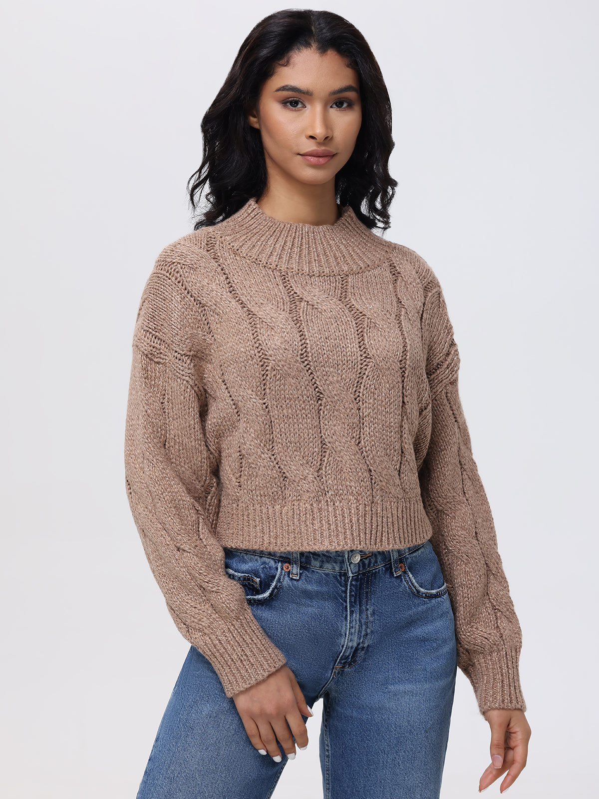 A woman with long dark hair poses against a plain backdrop in the 525 America Lola Lurex Cable-Knit Cropped Sweater. The textured beige fabric subtly shimmers with Lurex threading, perfectly paired with her blue jeans. Her hands rest casually at her sides.