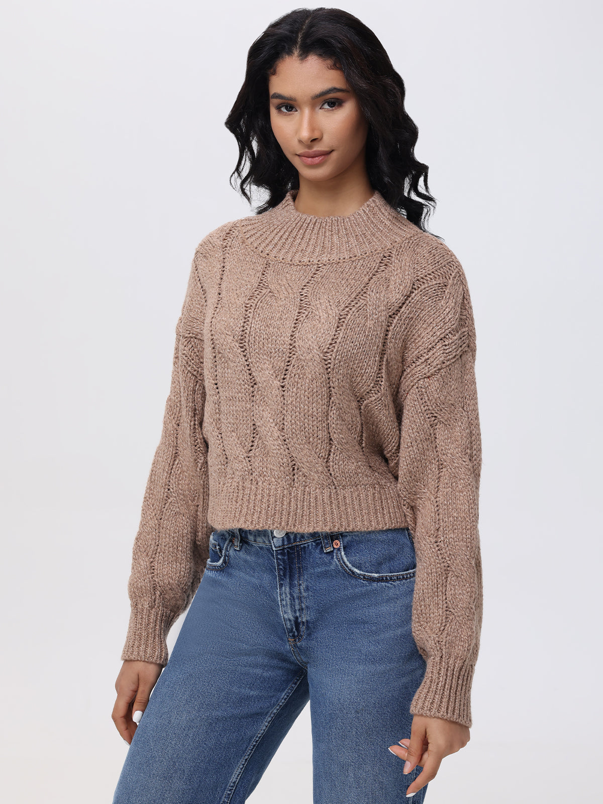 A person with long black hair stands against a plain backdrop in blue jeans and a 525 America Lola Lurex Cable-Knit Cropped Sweater, its intricate design subtly catching the light. They maintain a calm expression while looking directly at the camera.