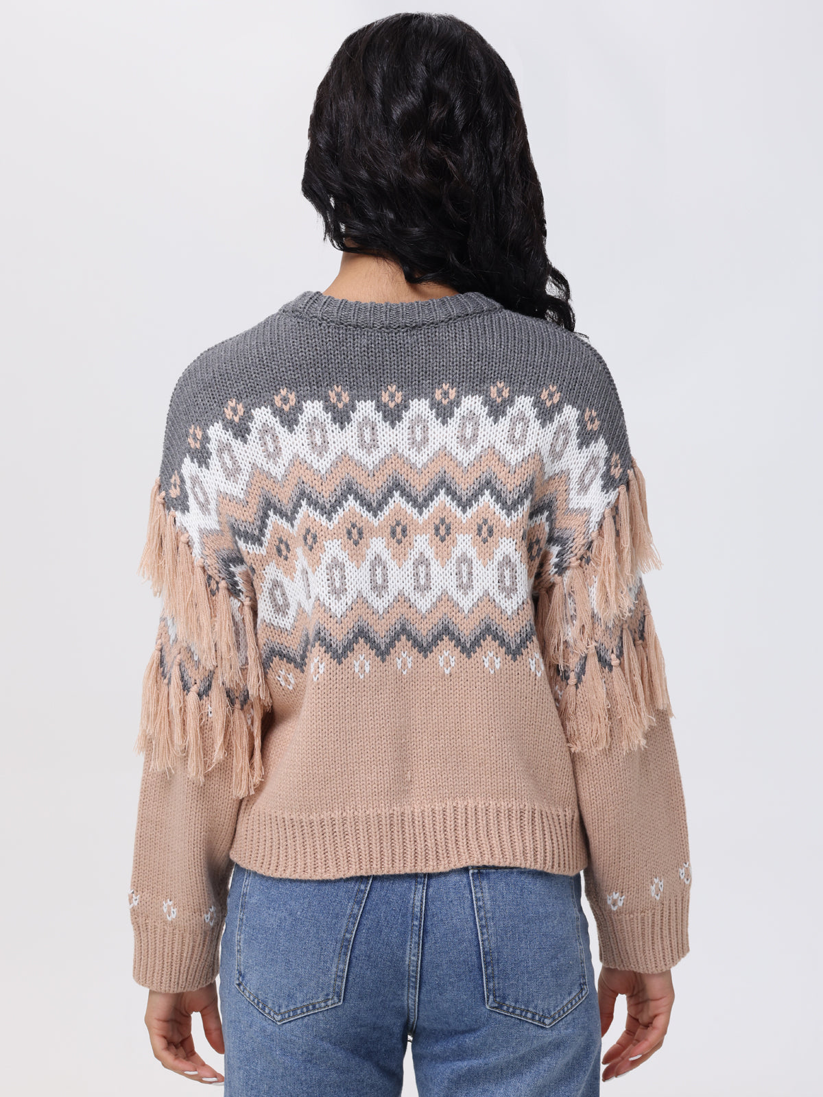 A person with long dark hair is wearing a 525 America relaxed-fit, multicolored knitted sweater featuring a geometric pattern and fringe details on the sleeves. Paired with blue jeans, they face away to showcase this winter essentials back against a plain background.