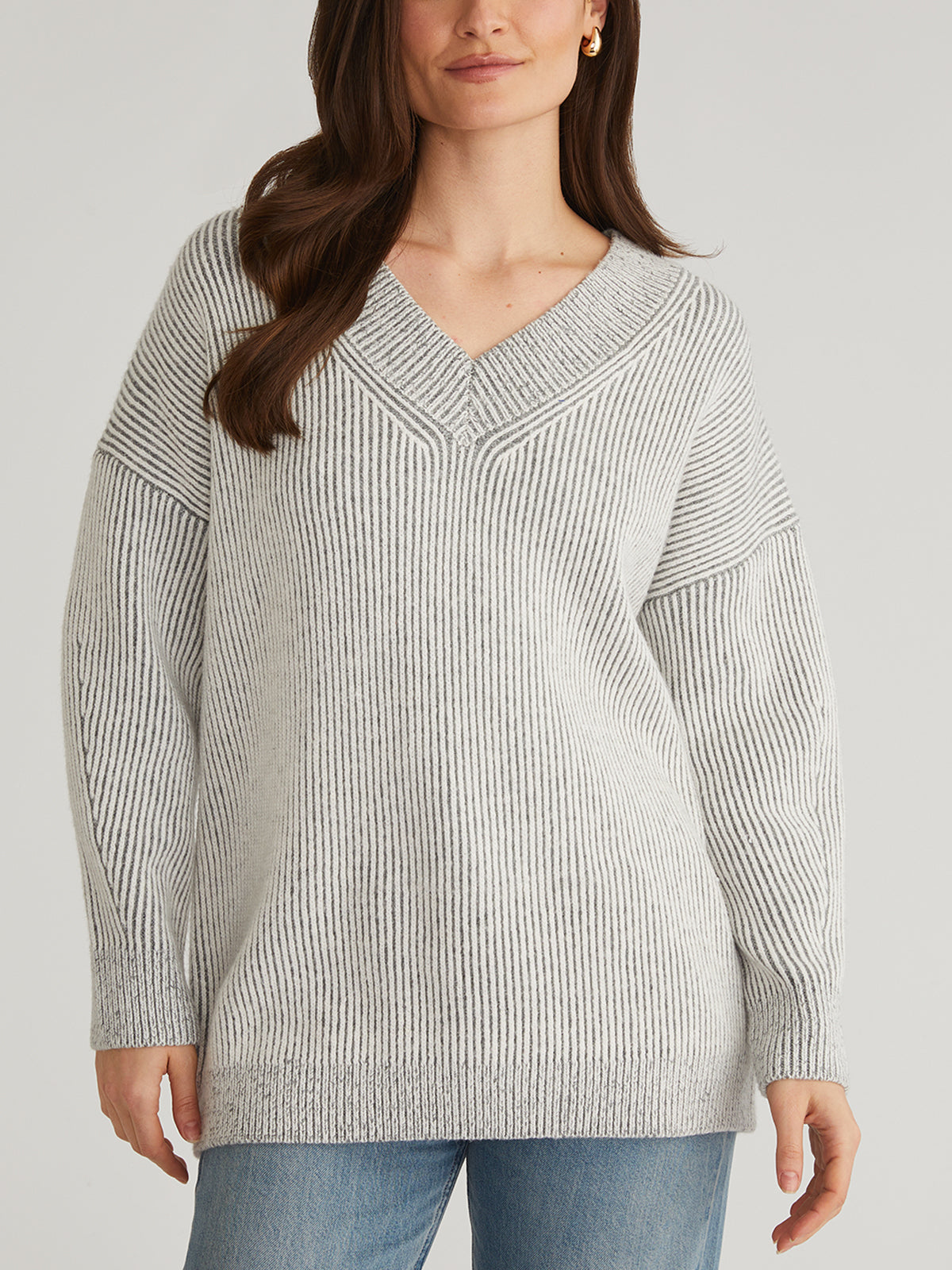 A person is wearing a stylish light gray V-neck sweater with long sleeves by 525 America, standing against a plain background. With long brown hair and turned slightly to the side, they pair the look with light blue jeans.