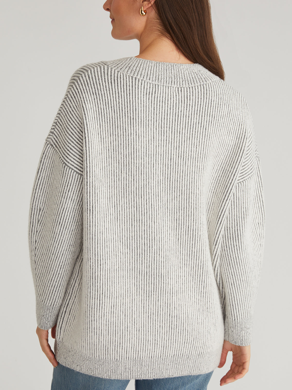 Wearing a 525 America gray contrast rib sweater with dropped shoulders and long sleeves, a fashion-forward individual stands with their back to us. Their brown hair flows over the V-neck, paired stylishly with blue jeans against an off-white background.