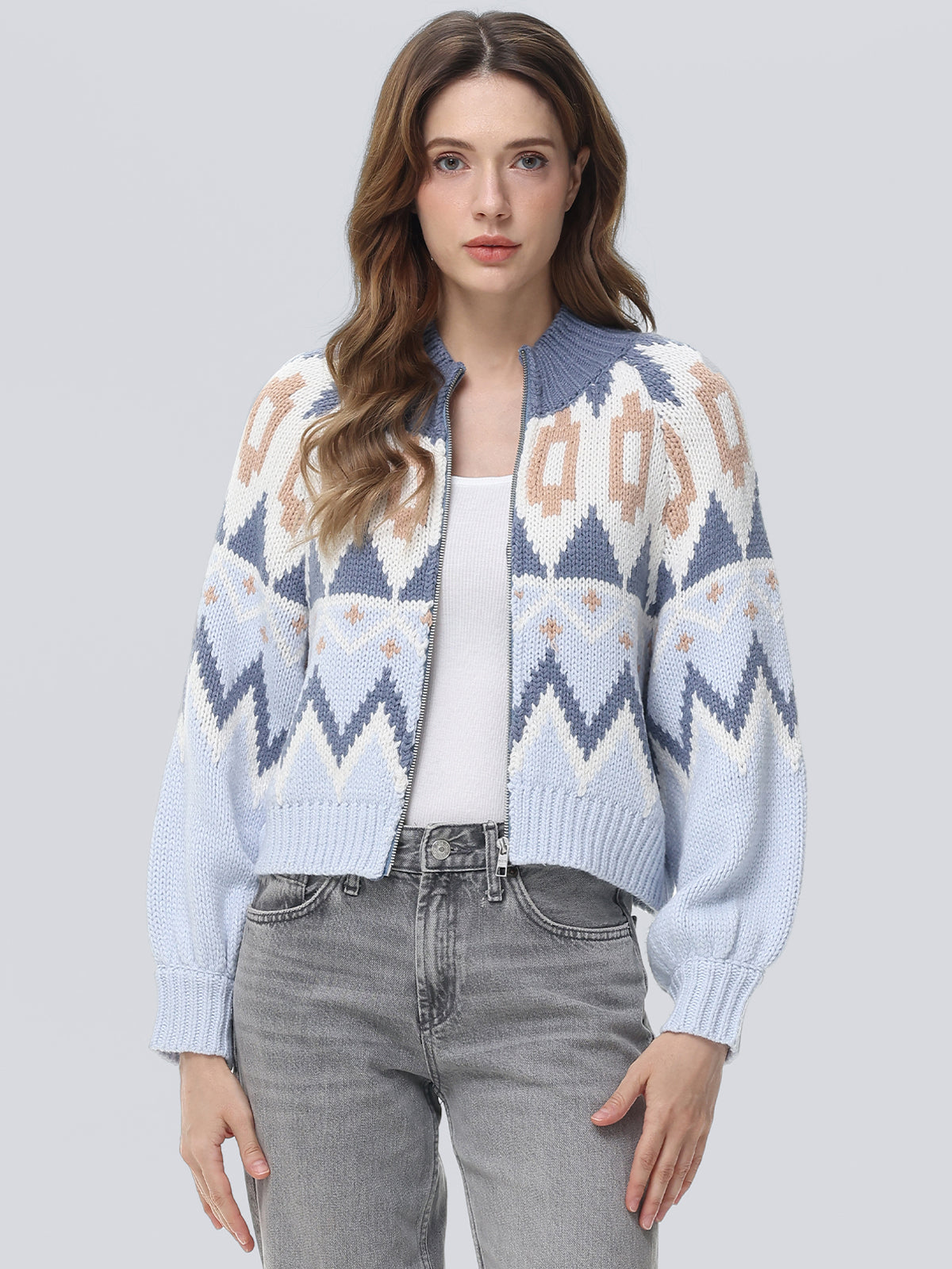 A person wearing the Victoria: Fair Isle Sweater Bomber Jacket with blue and white geometric winter patterns over a white top and gray jeans stands against a light gray background. The cardigan has diamond shapes, zigzags, and is made from 100% Acrylic for ultimate comfort.