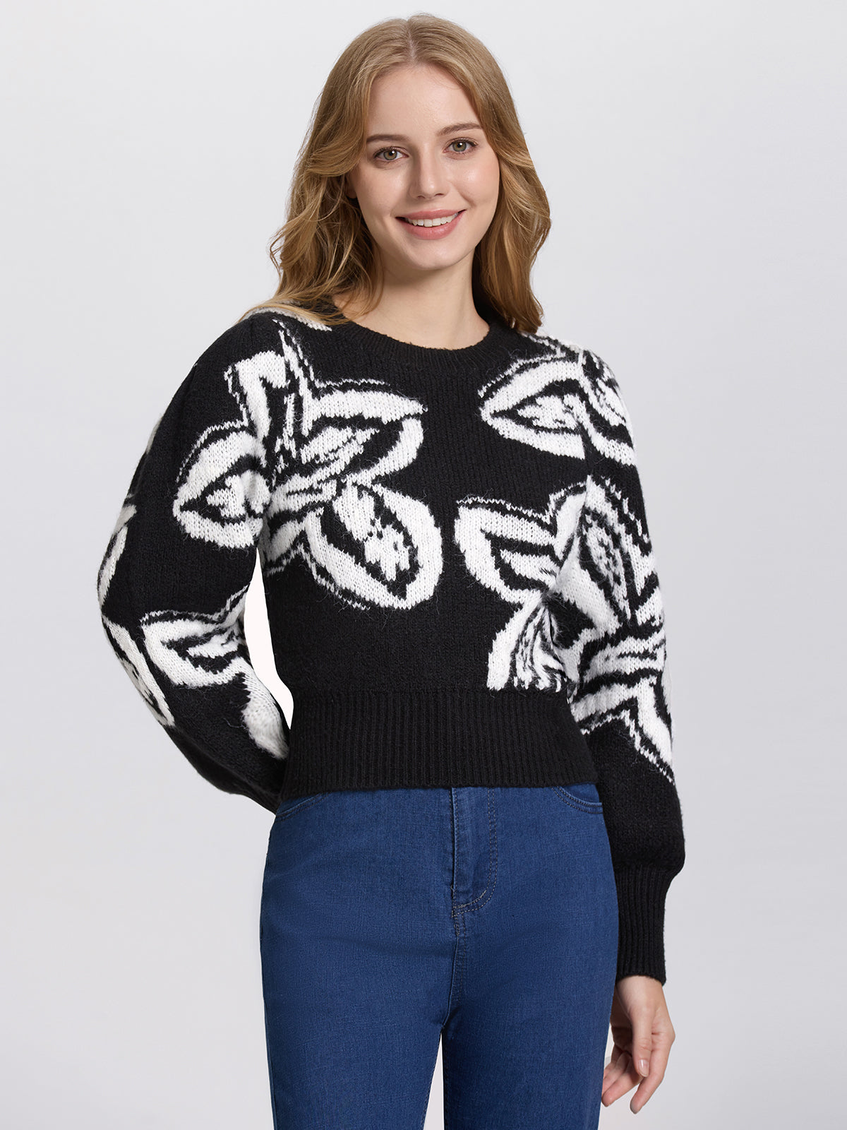 A woman with long, wavy hair wears a 525 America black cropped sweater with large white abstract patterns and blue jeans. She stands against a plain gray background, her crew neckline framing her face as she smiles warmly at the camera.