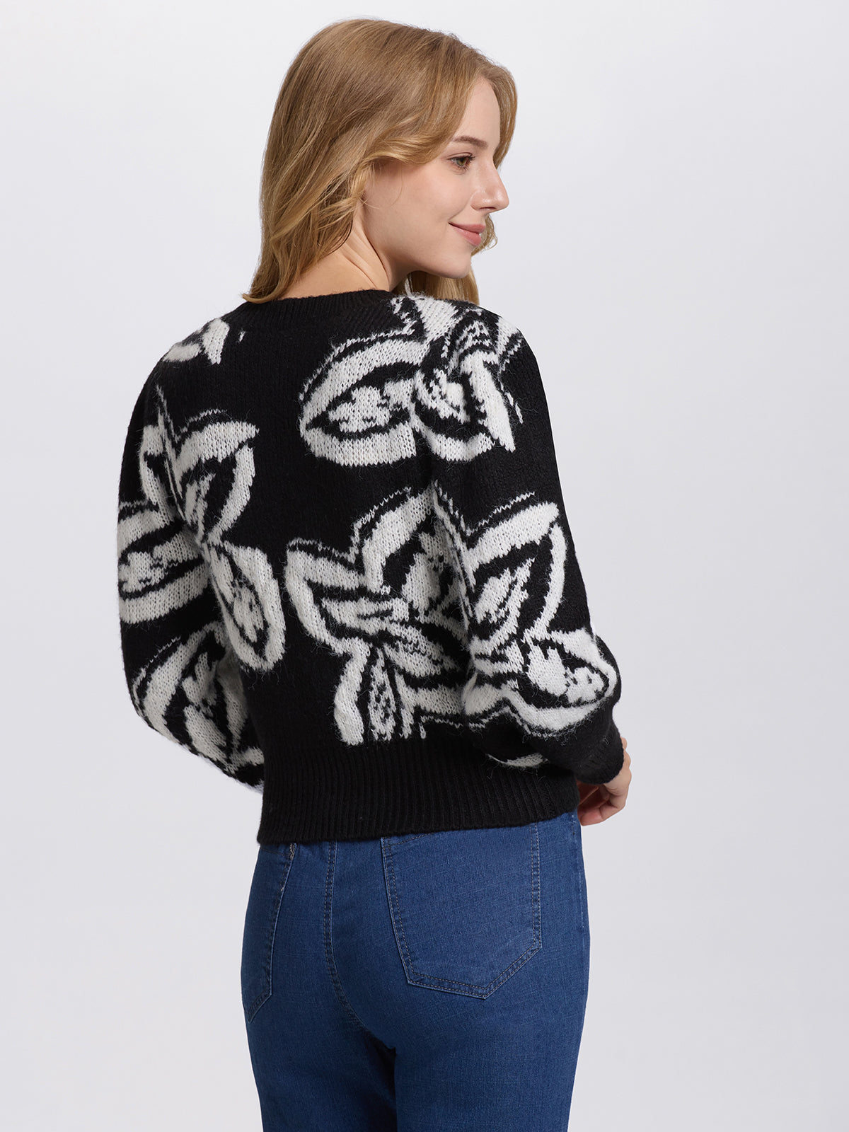 A person with long hair wears a 525 America black cropped sweater with large white floral jacquard patterns and blue jeans, featuring a crew neckline. They stand against a plain white background, looking to the left with a slight smile.