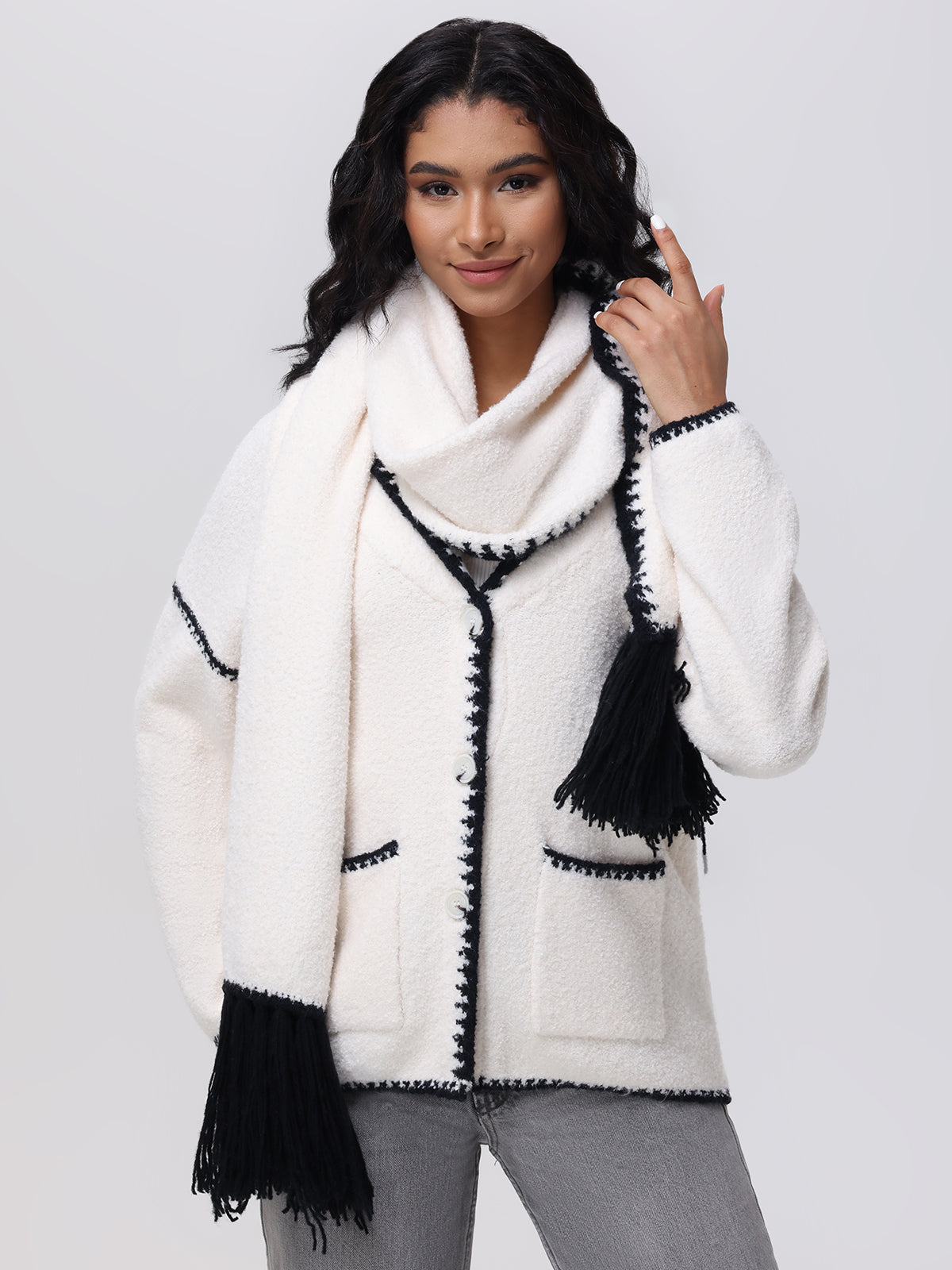 A person with wavy dark hair is wearing a structured white jacket from 525 America, showcasing whipstitch detailing and a removable scarf with black trim. Set against a plain backdrop, they gracefully hold the scarf and pair it with gray pants for a modern look.