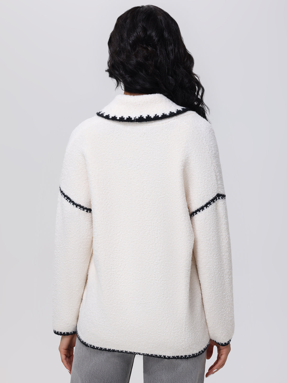 A person with long dark hair is wearing a white fleece jacket with black trim from 525 America. The textured design, similar to the Tierney Whipstitch Cardigan, features decorative edges at the collar, shoulders, and sleeve hems. The individual faces away against a plain background.