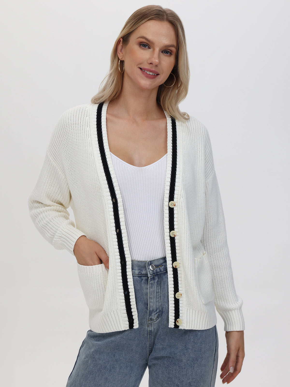 A person dons a 525 America New York Cardigan made of 100% cotton, featuring a relaxed fit, black trim, and gold buttons. They stand against a plain background with one hand in their pocket, paired with a white top and blue jeans.